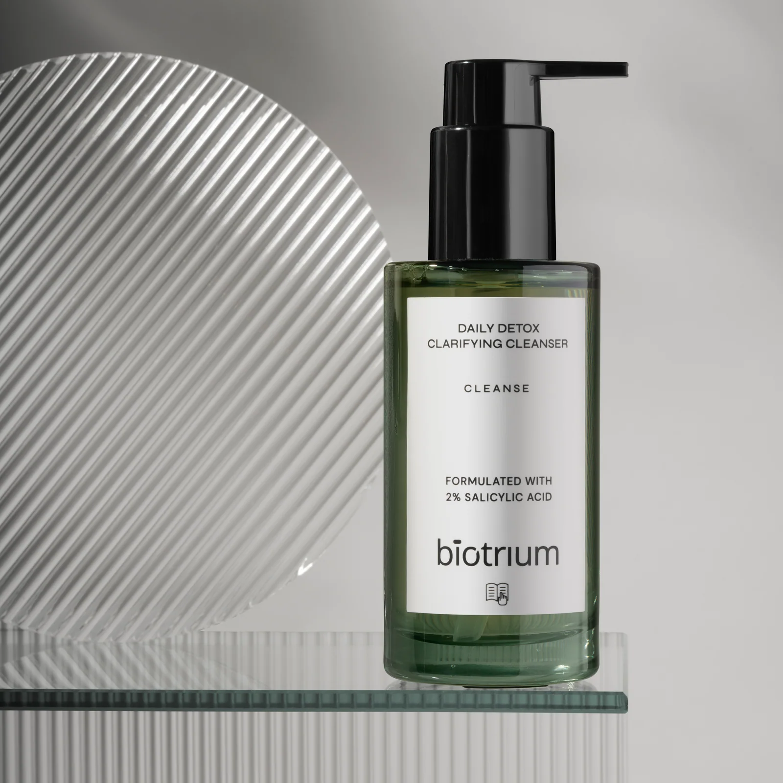 Green Biotrium Daily Detox cleanser bottle on a glass shelf beside a circular ribbed-glass panel.