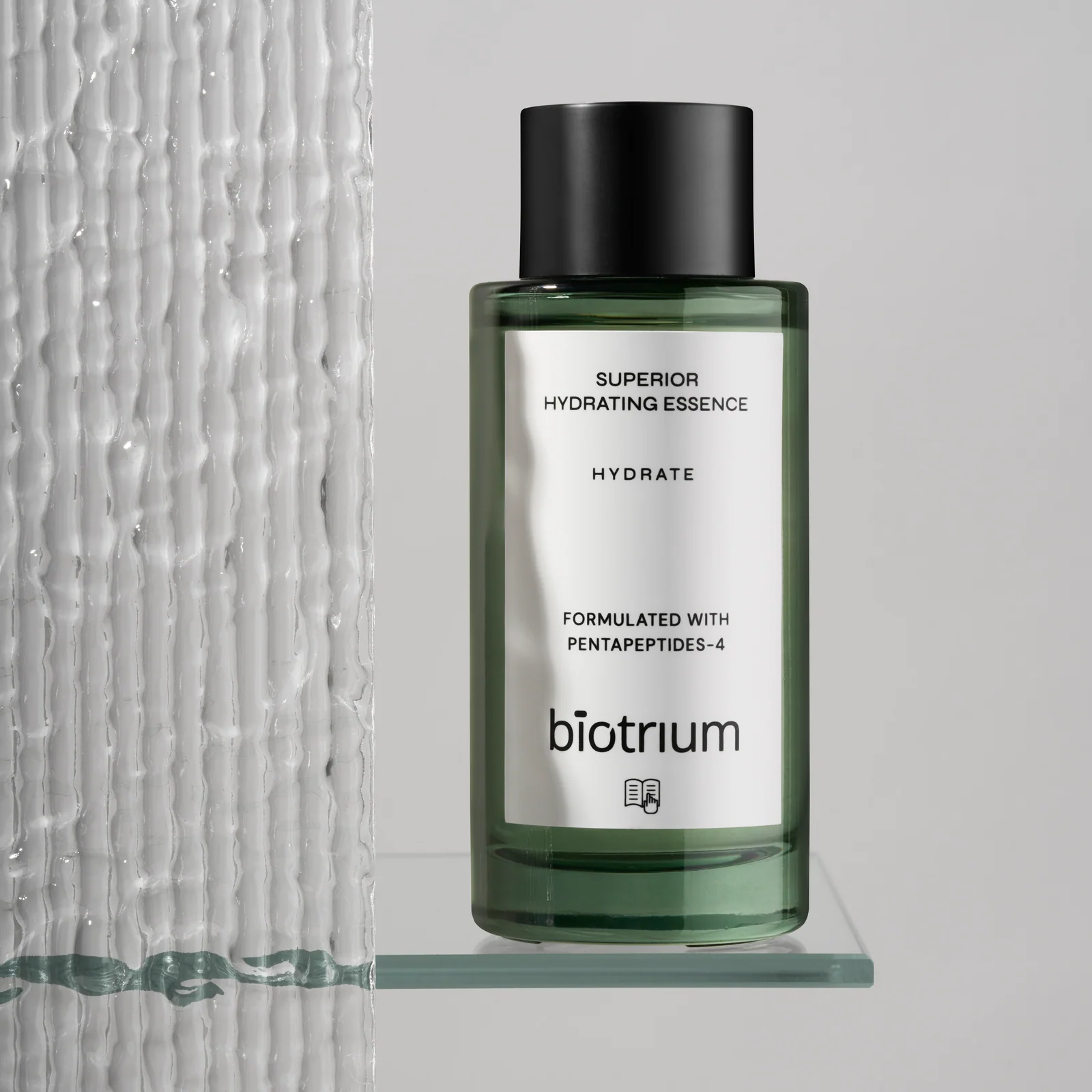 Green Biotrium Hydrating Essence bottle with a black cap on a glass shelf beside textured glass.