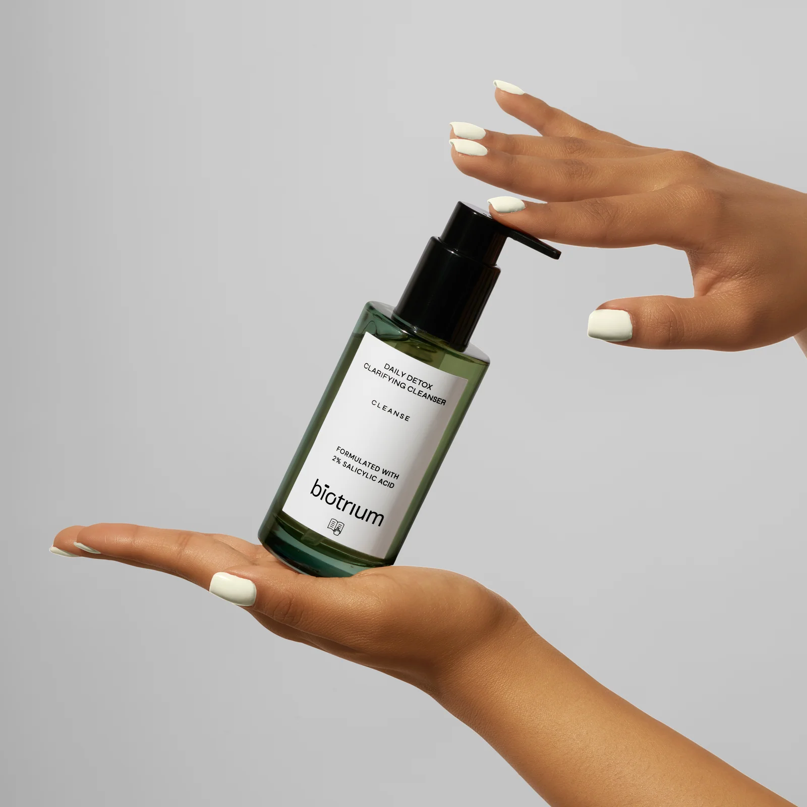 Hands holding a green Biotrium cleanser bottle with a finger resting on its black pump against grey.