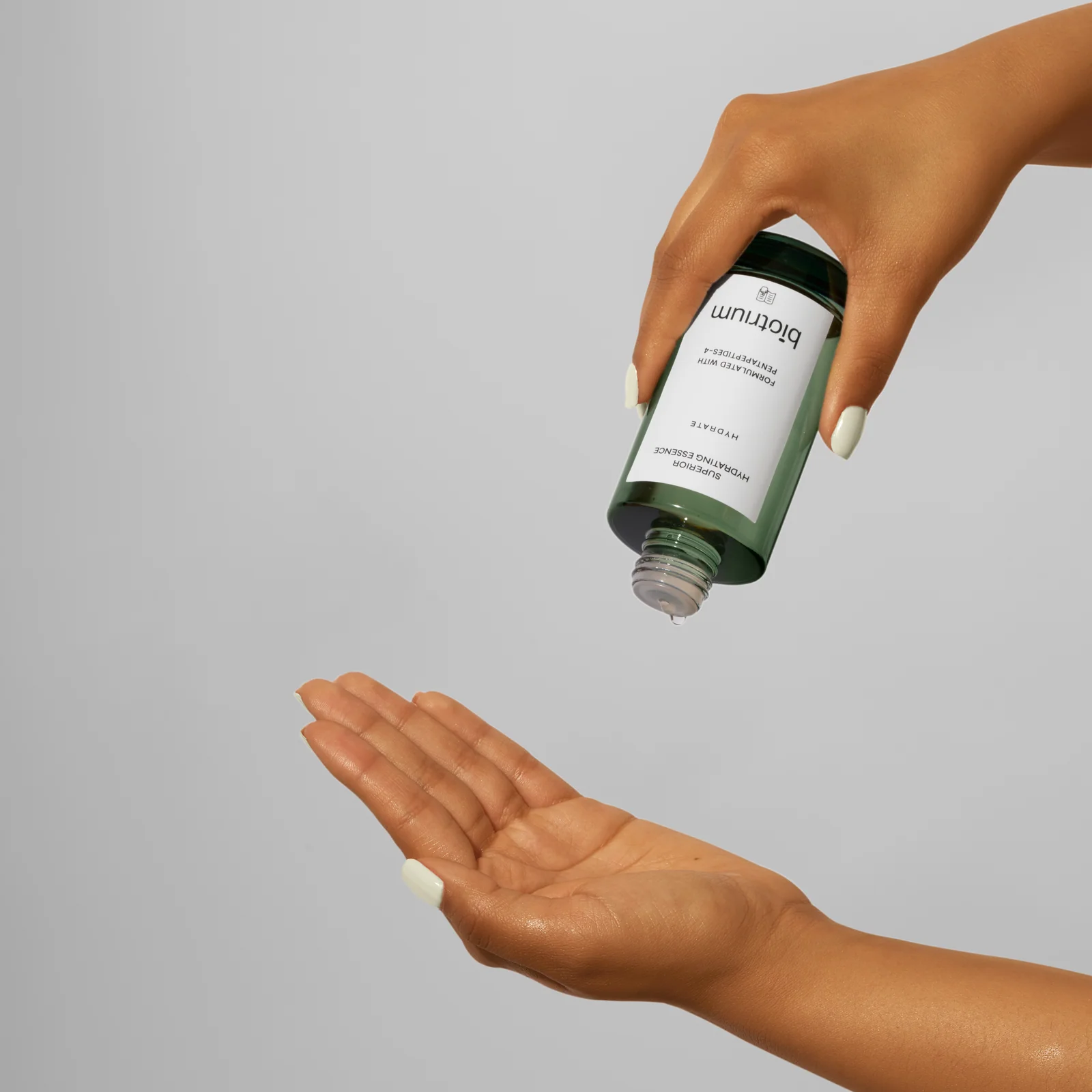 Hand tilting an open green Biotrium Hydrating Essence bottle above an outstretched palm against grey.