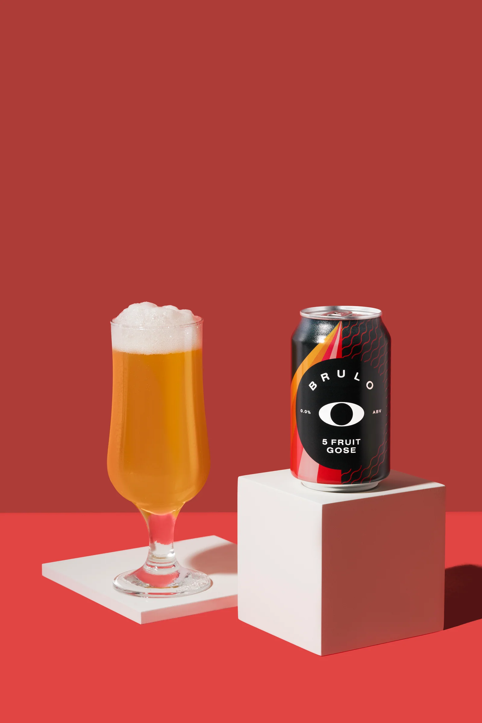 Brülo 5 Fruit Gose can and a filled beer glass on white platforms against red.