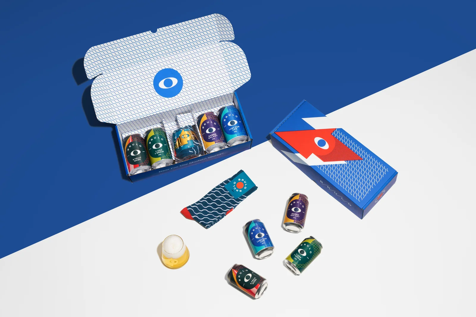 Open Brülo gift box with beer cans, a glass and socks arranged across a blue-and-white surface.