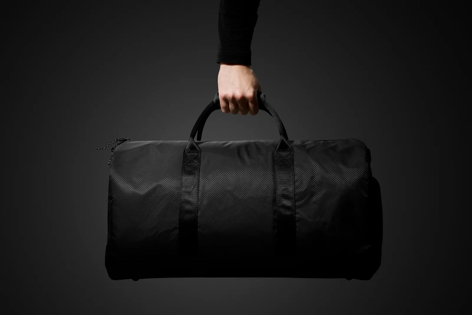 Hand holding a black Double Oak duffel bag by its handles against dark grey.