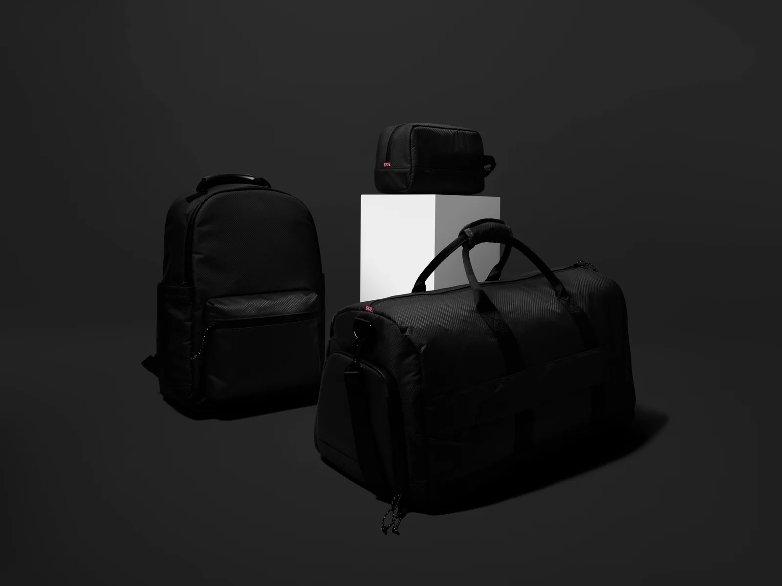 Black Double Oak backpack, duffel bag and zip pouch arranged around a white plinth against dark grey.