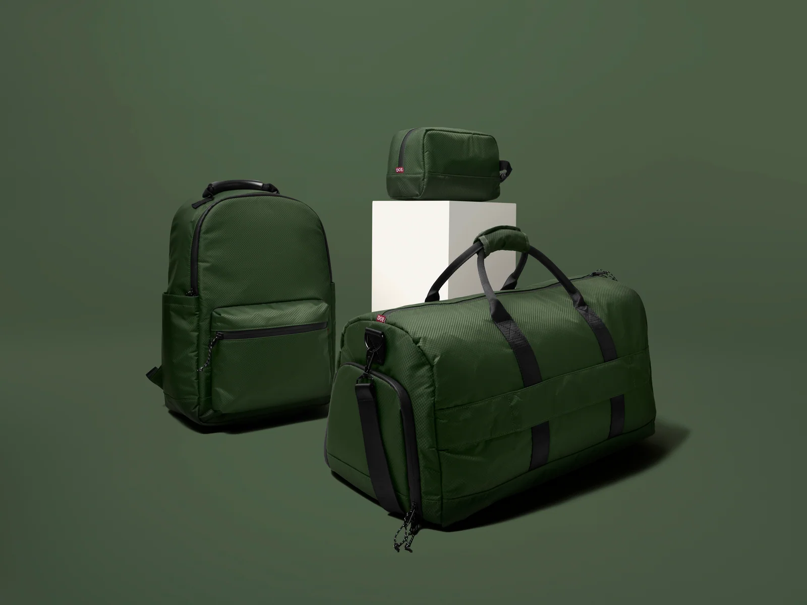 Green Double Oak backpack, duffel bag and zip pouch arranged around a white plinth against green.
