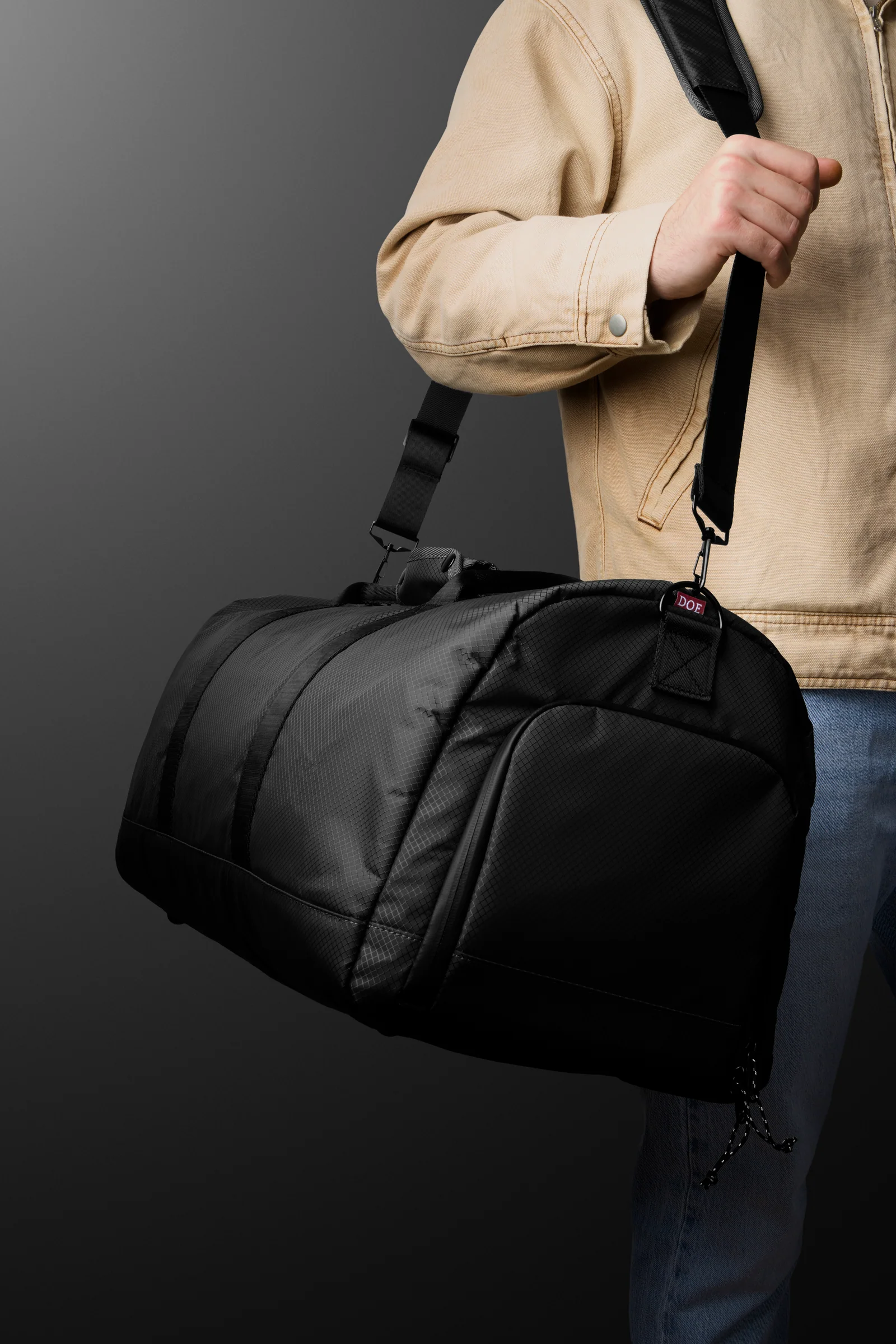 Person in a beige jacket carrying a black Double Oak duffel bag on a shoulder strap.