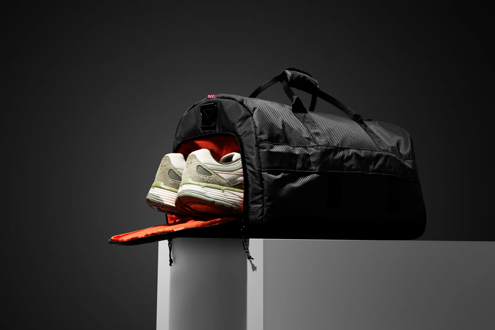 Black Double Oak duffel bag with its end compartment open and trainers visible on a grey plinth.