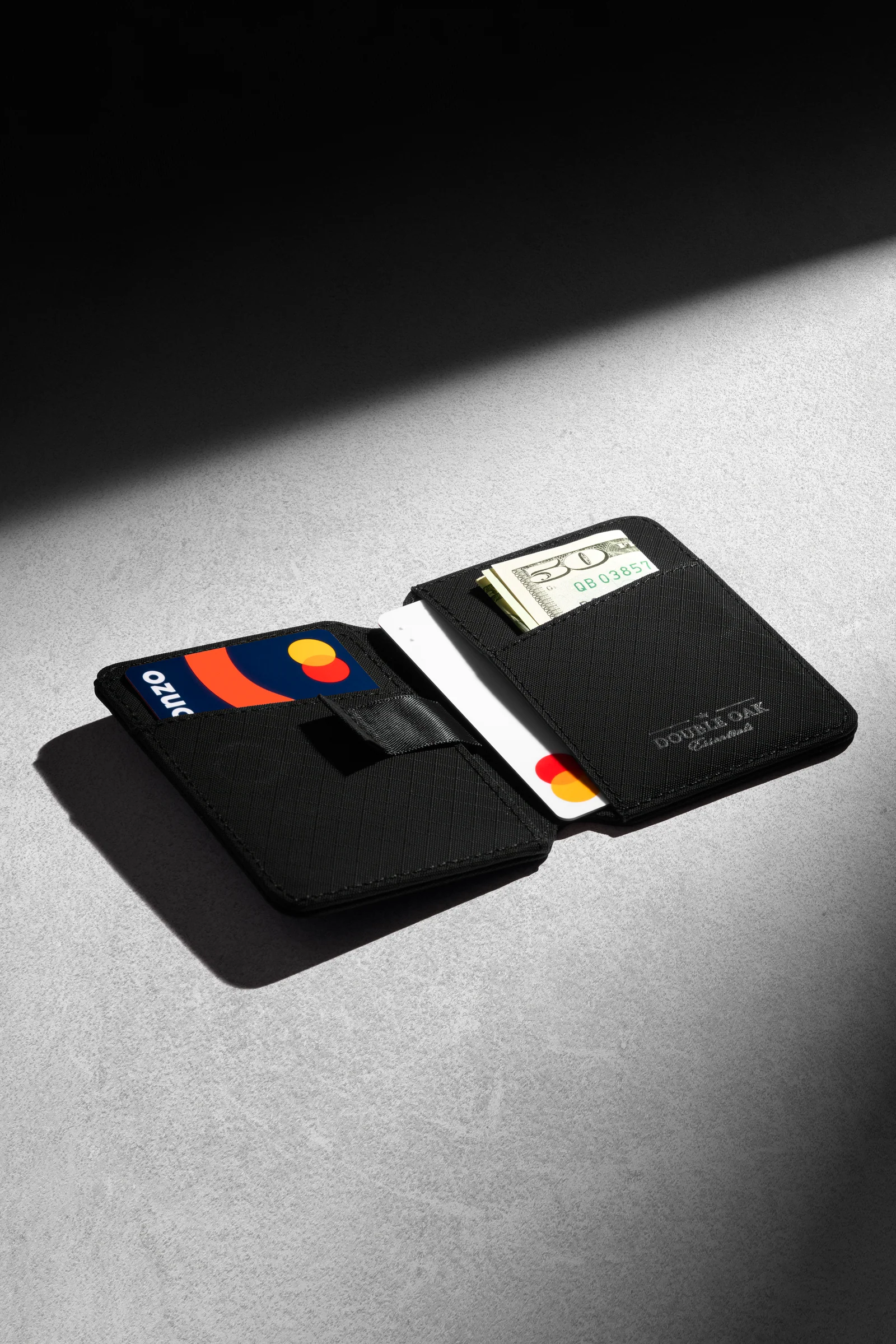 Open black Double Oak wallet with cards and a banknote on grey in directional light.