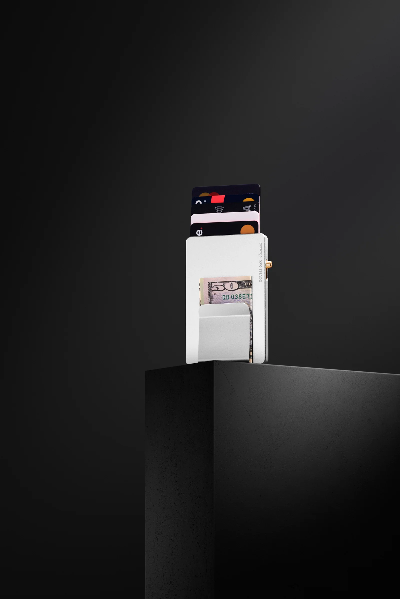 Silver-coloured Double Oak cardholder with raised cards and a banknote on a dark plinth.