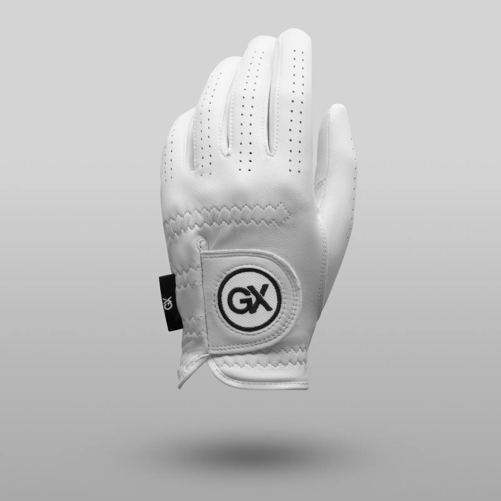 Angled rear view of a white GX Golf glove with perforated fingers and a round logo patch above a soft shadow on grey.