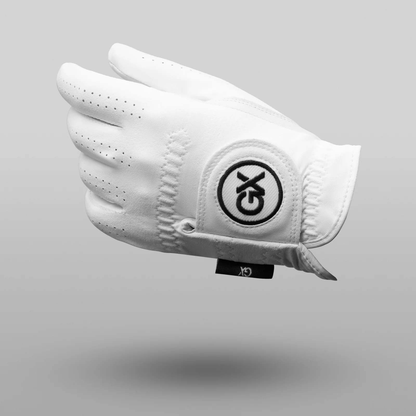 Curved white GX Golf glove with perforated fingers and a round logo patch shown from the back above a soft shadow on grey.