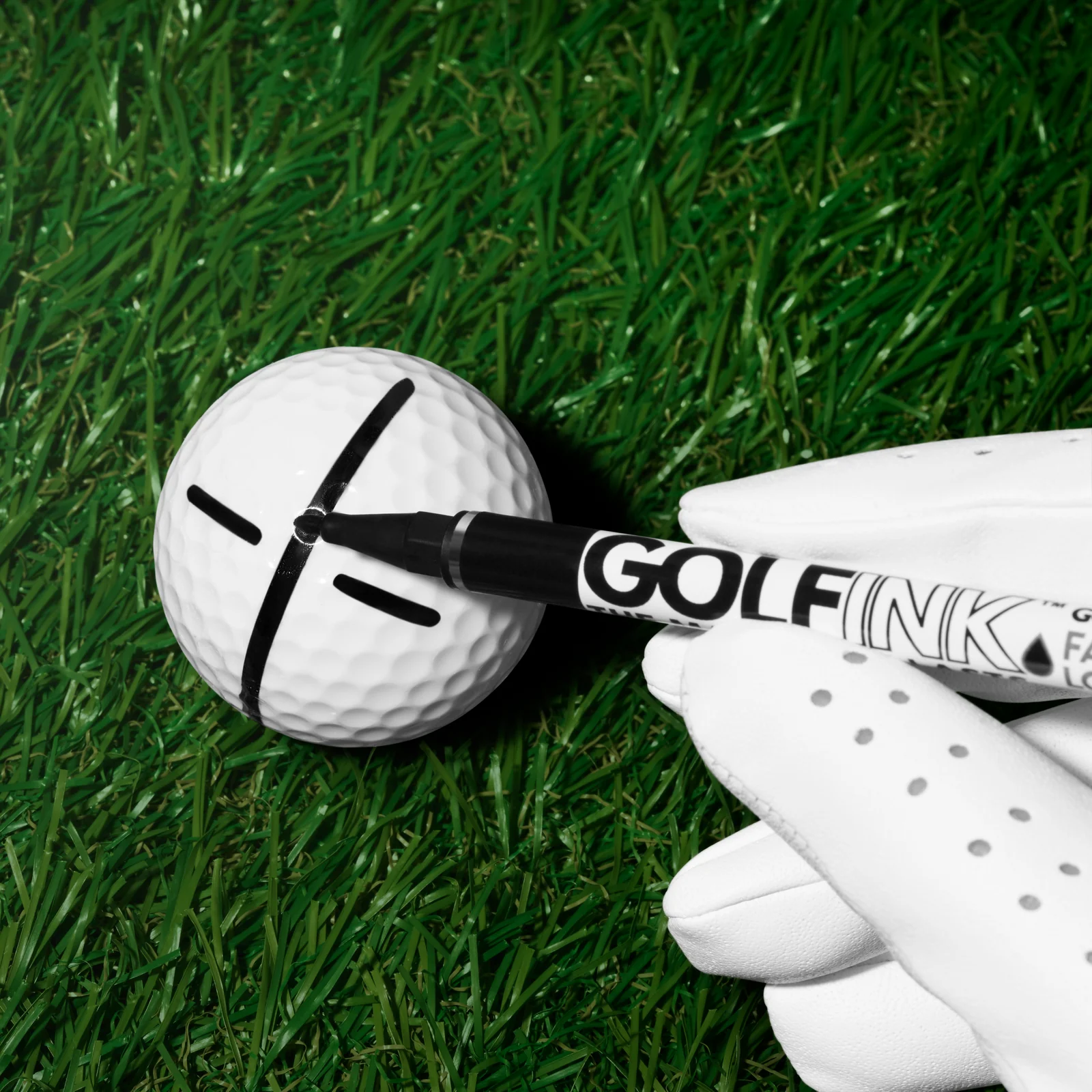 White-gloved hand using a black Golf Ink pen to mark a golf ball on grass.