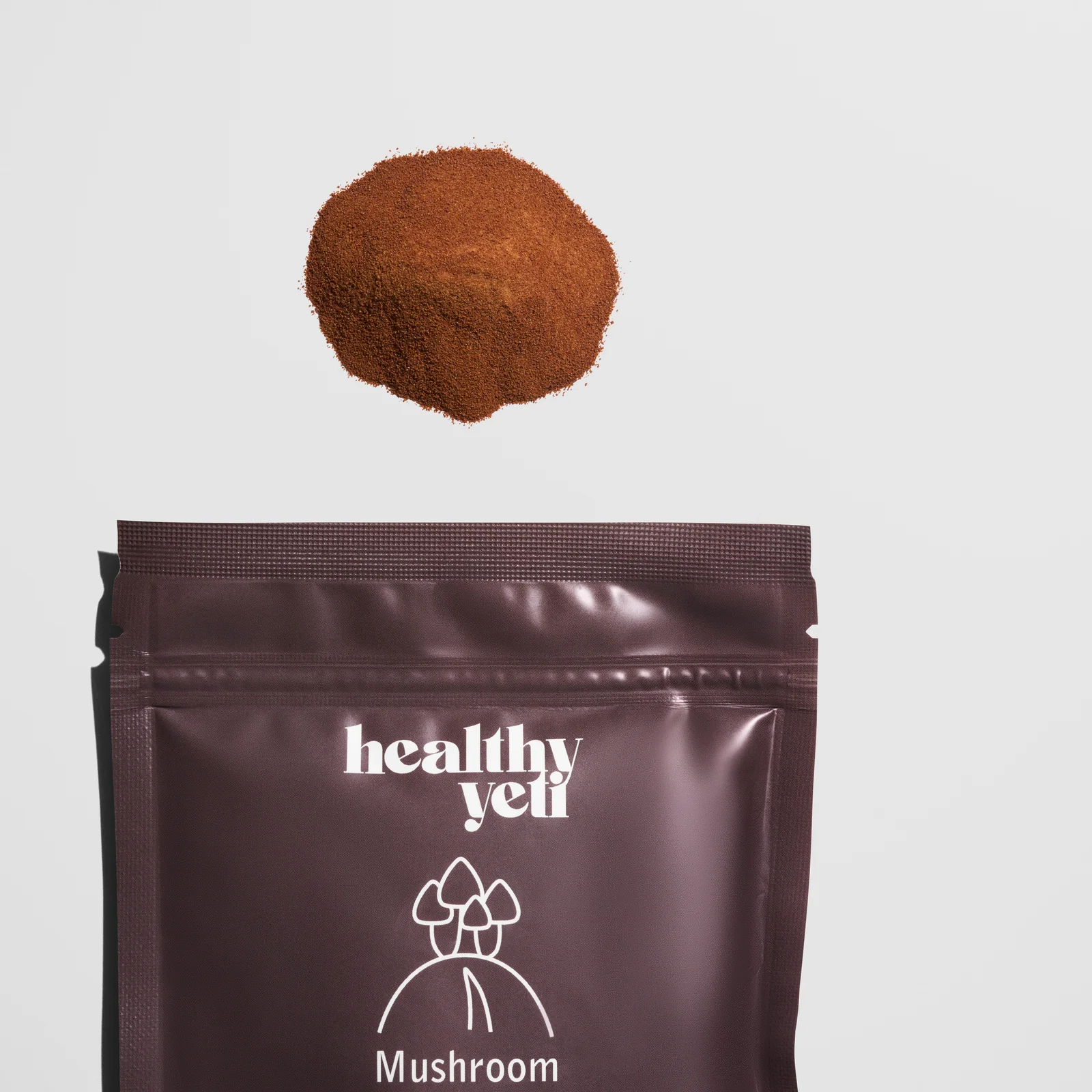 Mound of brown powder suspended above a cropped Healthy Yeti Mushroom Coffee pouch on a light background.