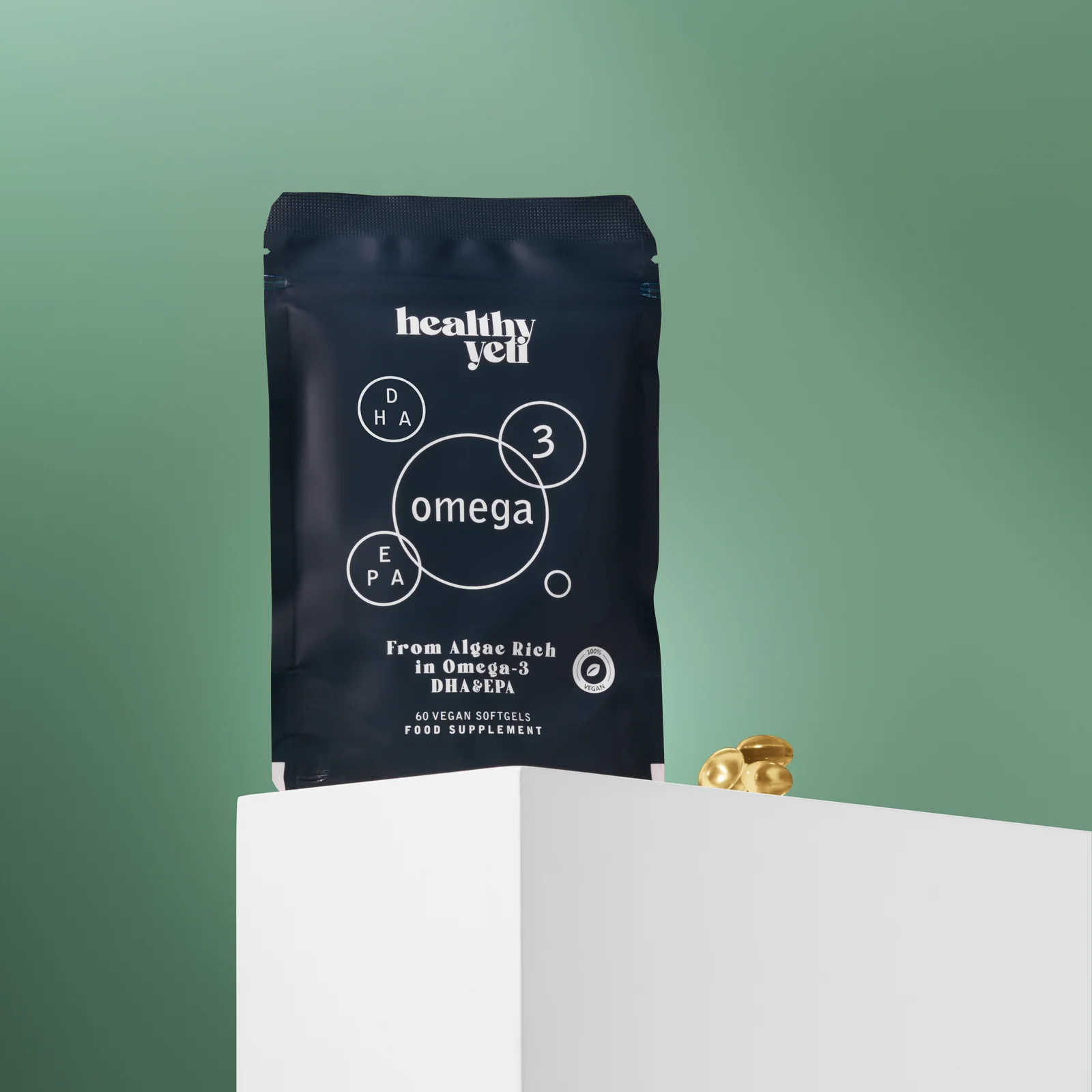 Blue Healthy Yeti Omega 3 pouch and golden capsules behind a white plinth against green.