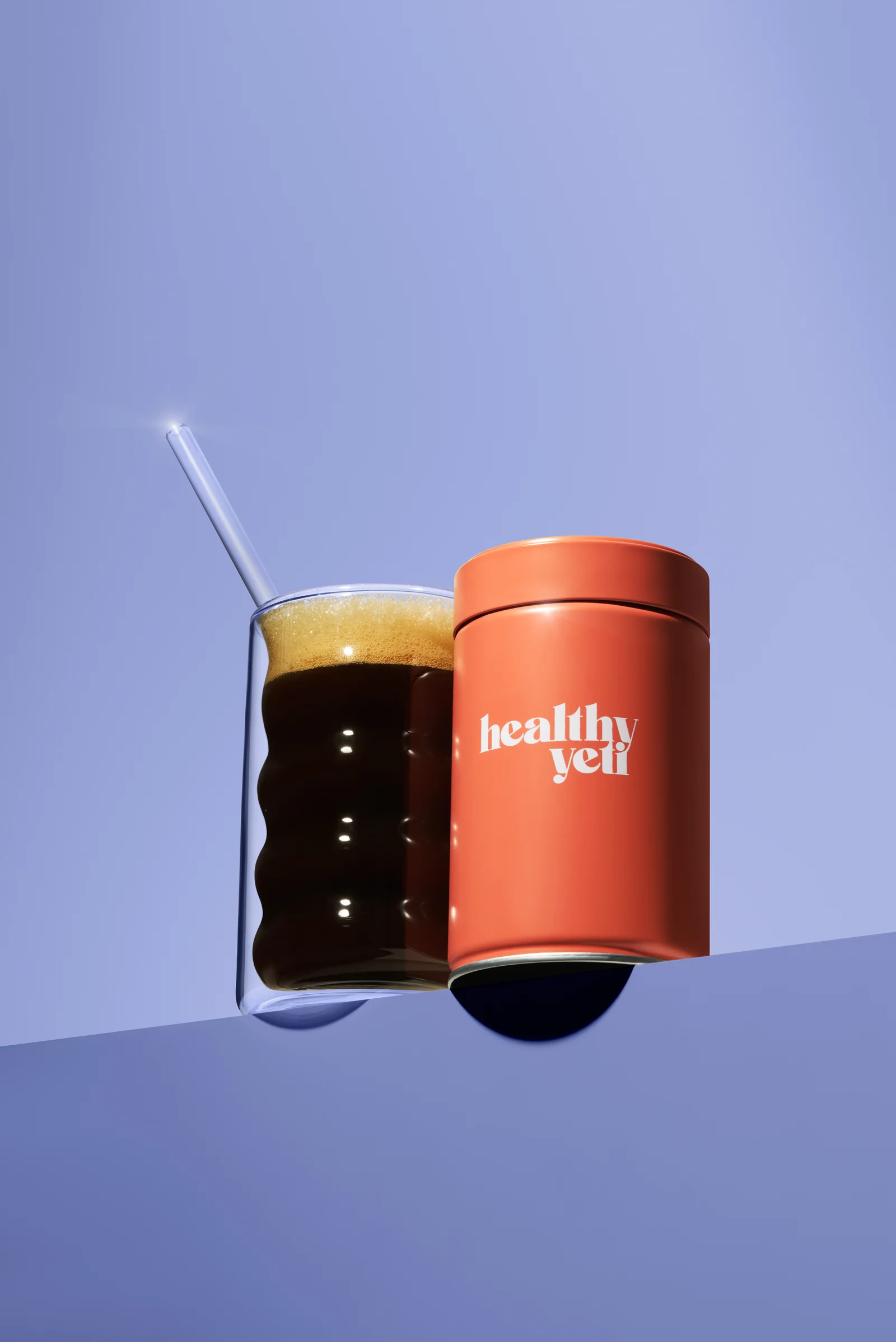Orange Healthy Yeti jar beside a glass of dark coffee against lilac.