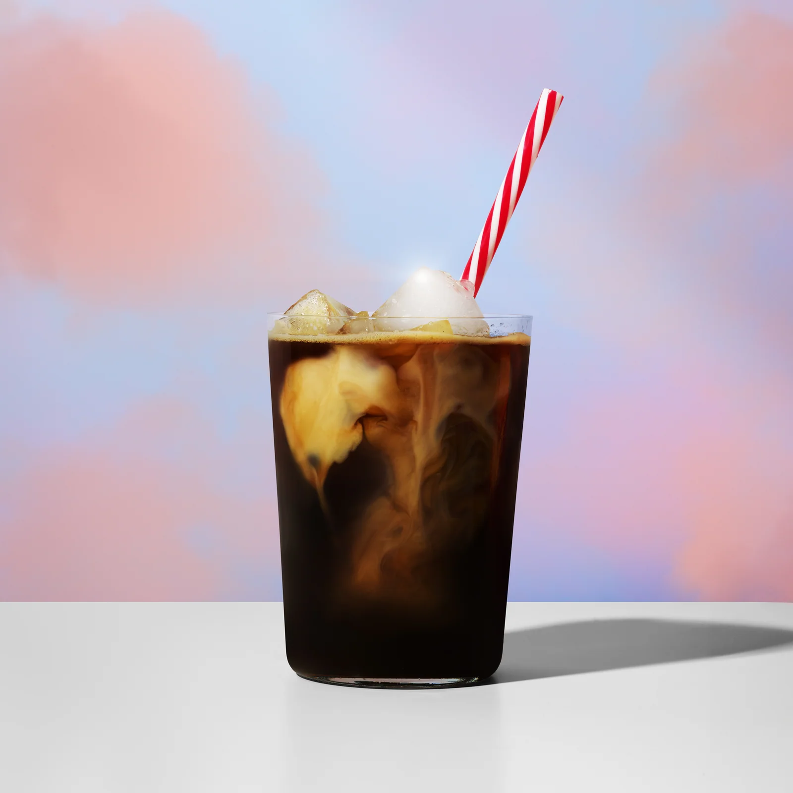 Glass of iced coffee with swirling milk and a red-and-white straw against a pastel sky.