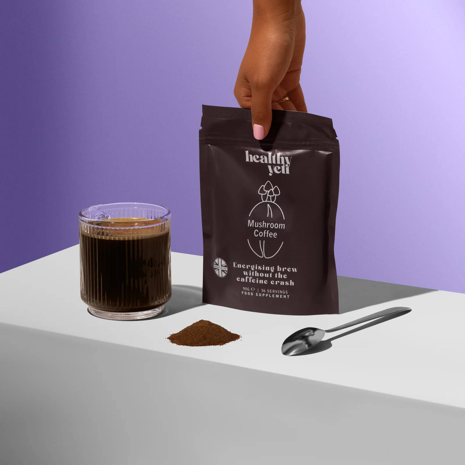 Hand holding a Healthy Yeti Mushroom Coffee pouch beside a glass of coffee, powder and a scoop on a white plinth.