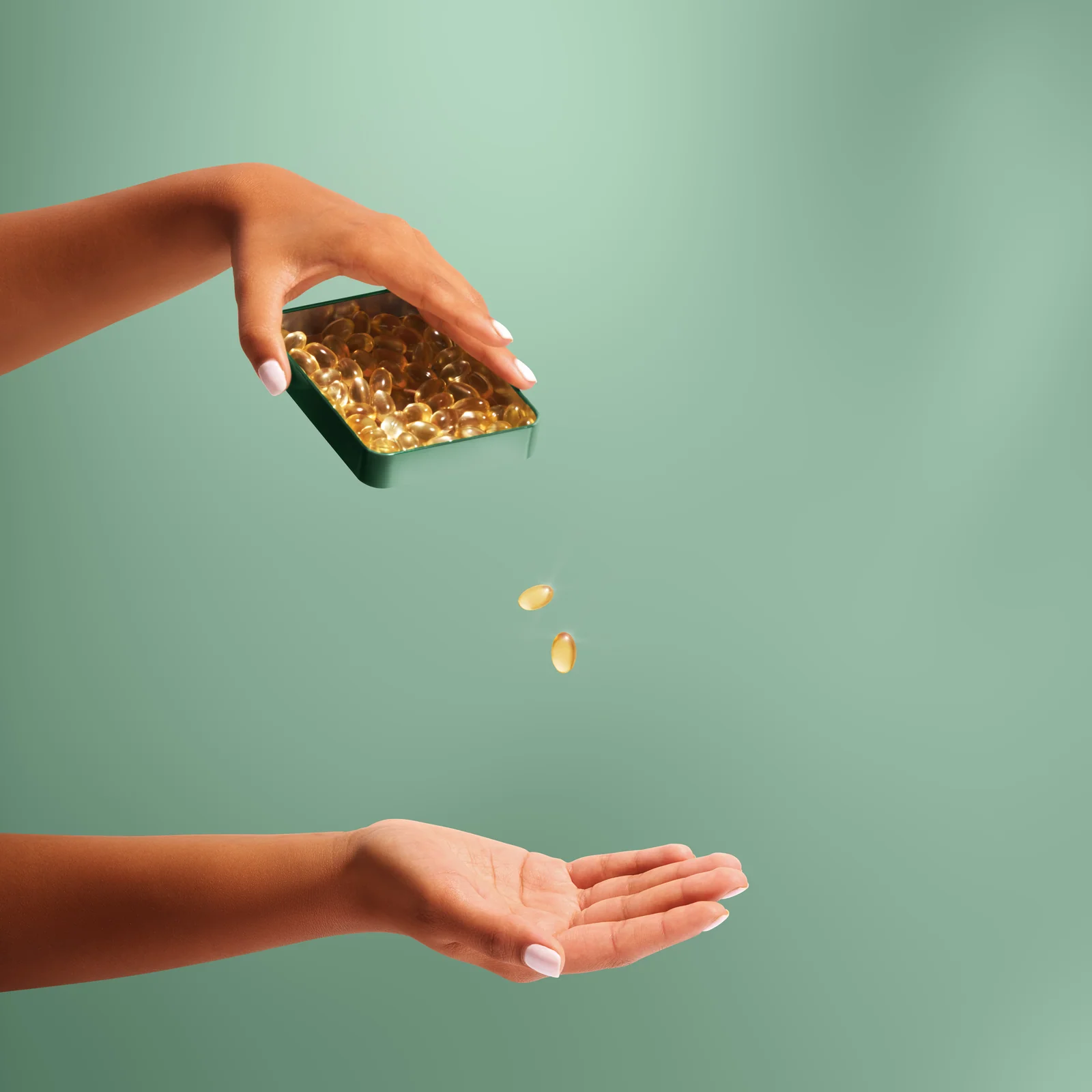 Hands tipping golden capsules from a green tin towards an open palm against green.