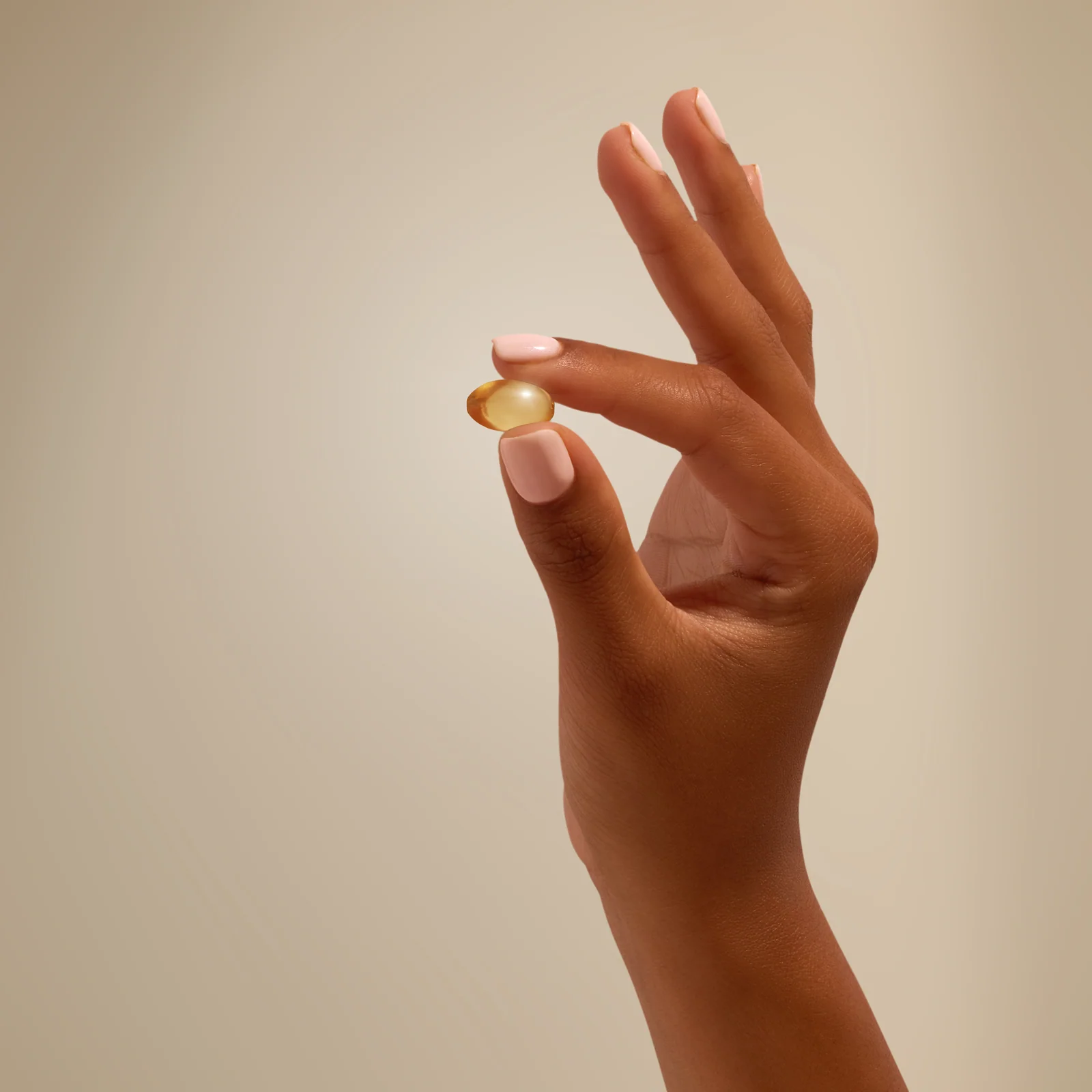 Hand holding a golden capsule between finger and thumb against beige.