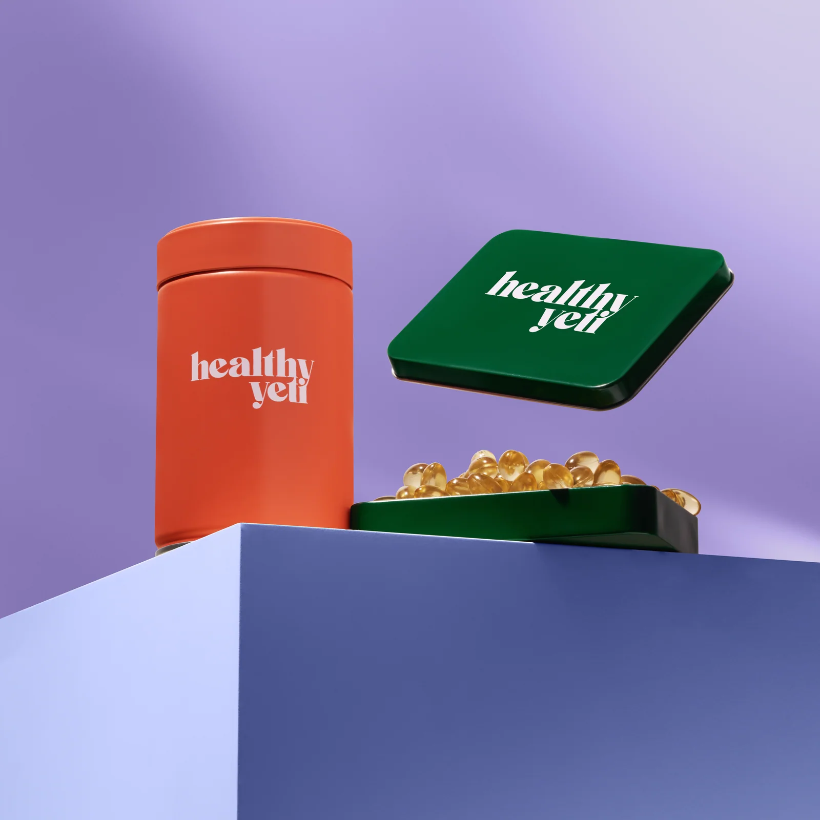 Orange Healthy Yeti jar beside an open green tin of golden capsules on a lilac plinth.