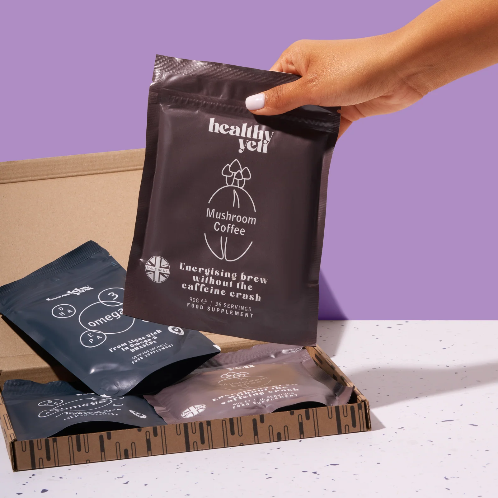 Hand lifting a Healthy Yeti Mushroom Coffee pouch from an open delivery box against lilac.