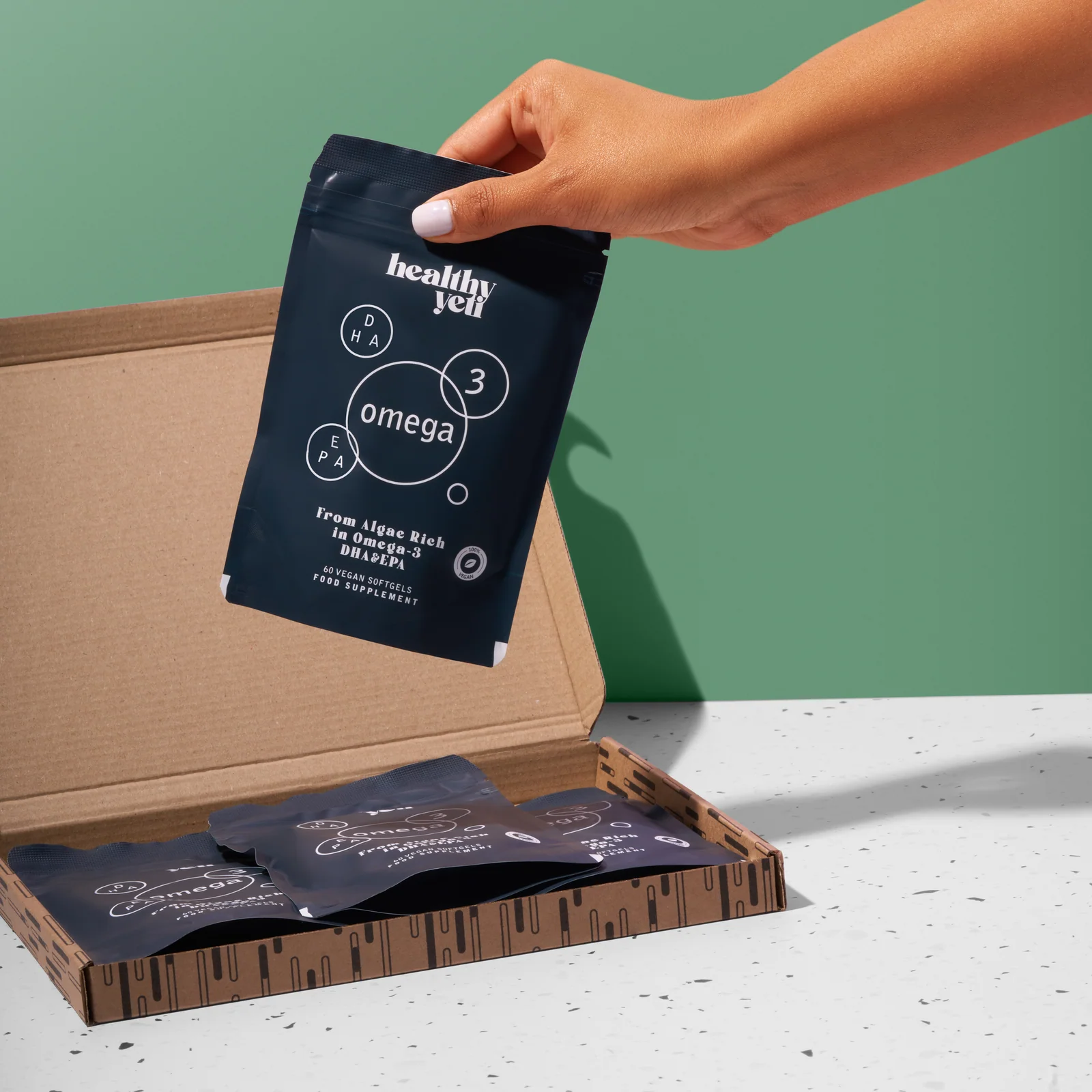 Hand lifting a Healthy Yeti Omega 3 pouch from an open delivery box against green.