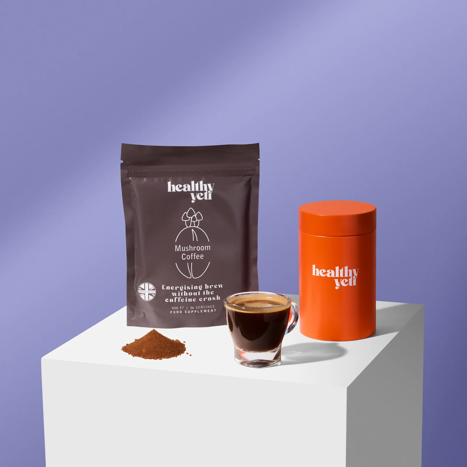 Healthy Yeti Mushroom Coffee pouch, orange tin, espresso and powder on a white plinth against lilac.