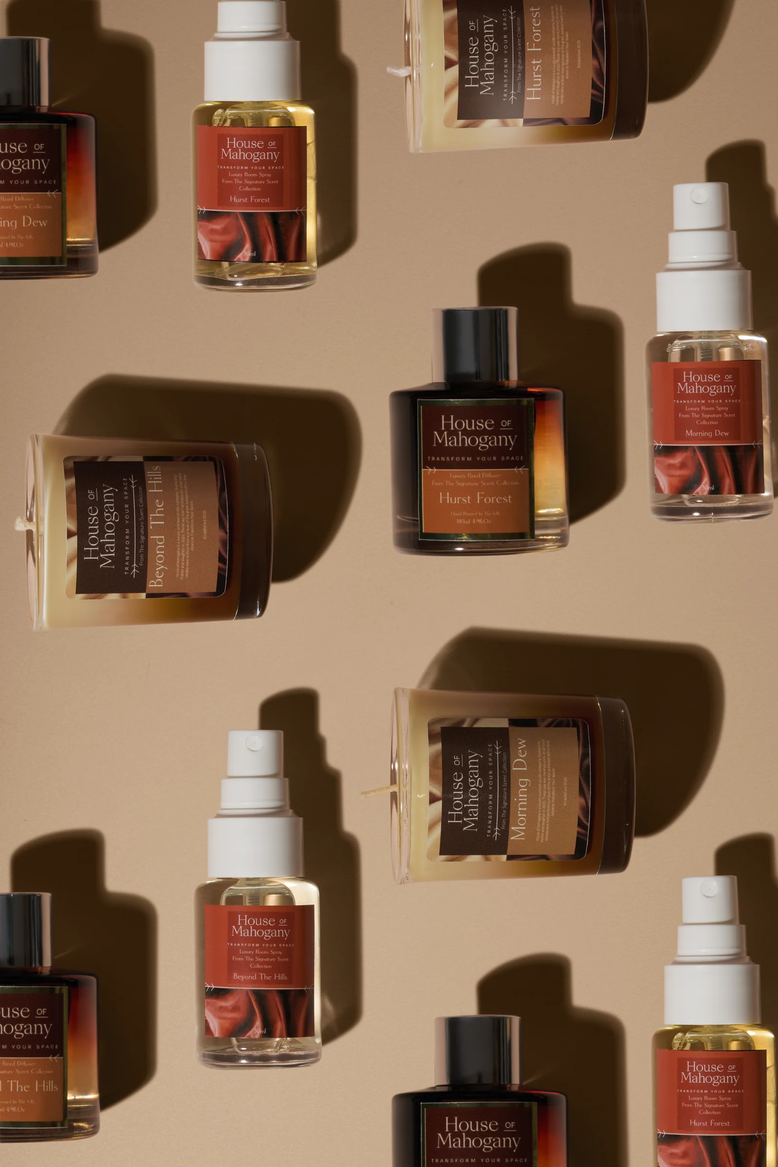 House of Mahogany candles, room sprays and fragrance bottles arranged in a repeating pattern on brown with strong shadows.