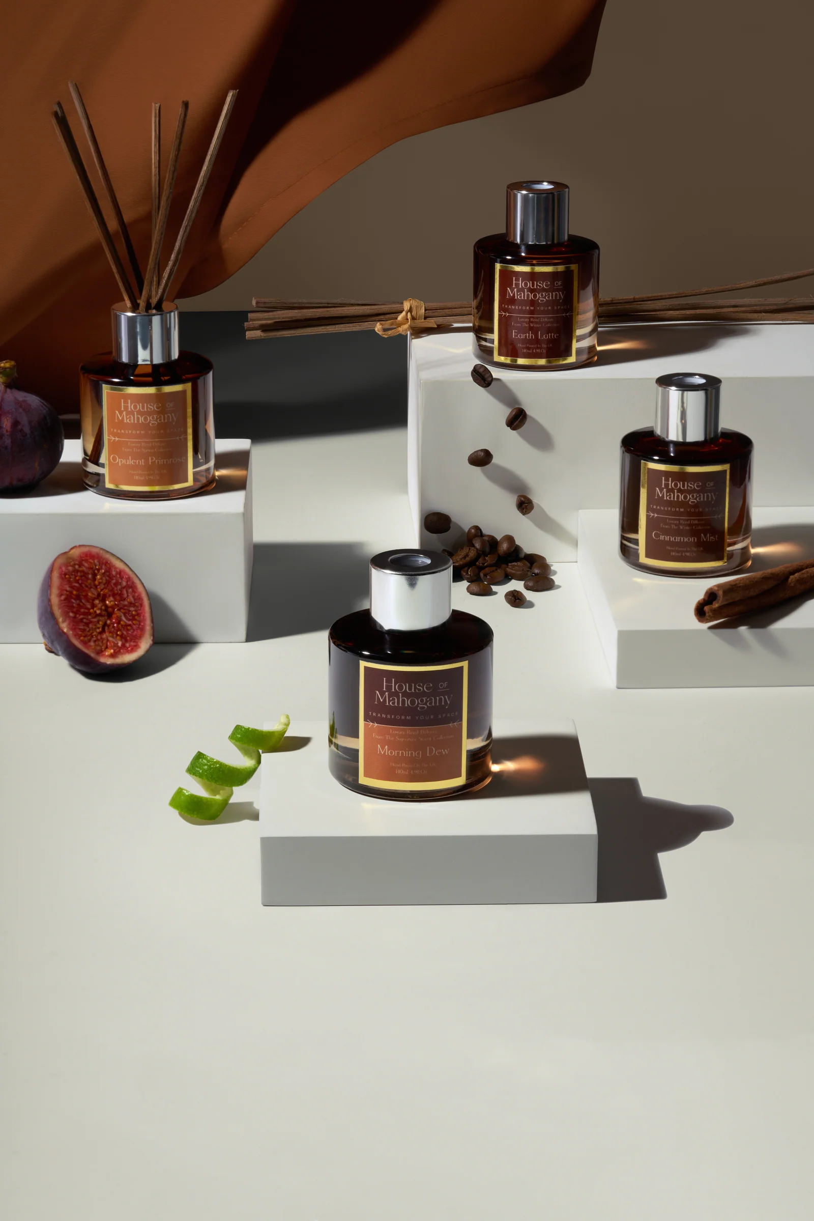 House of Mahogany fragrance bottles and a reed diffuser on stepped cream plinths with fig, coffee beans and cinnamon.