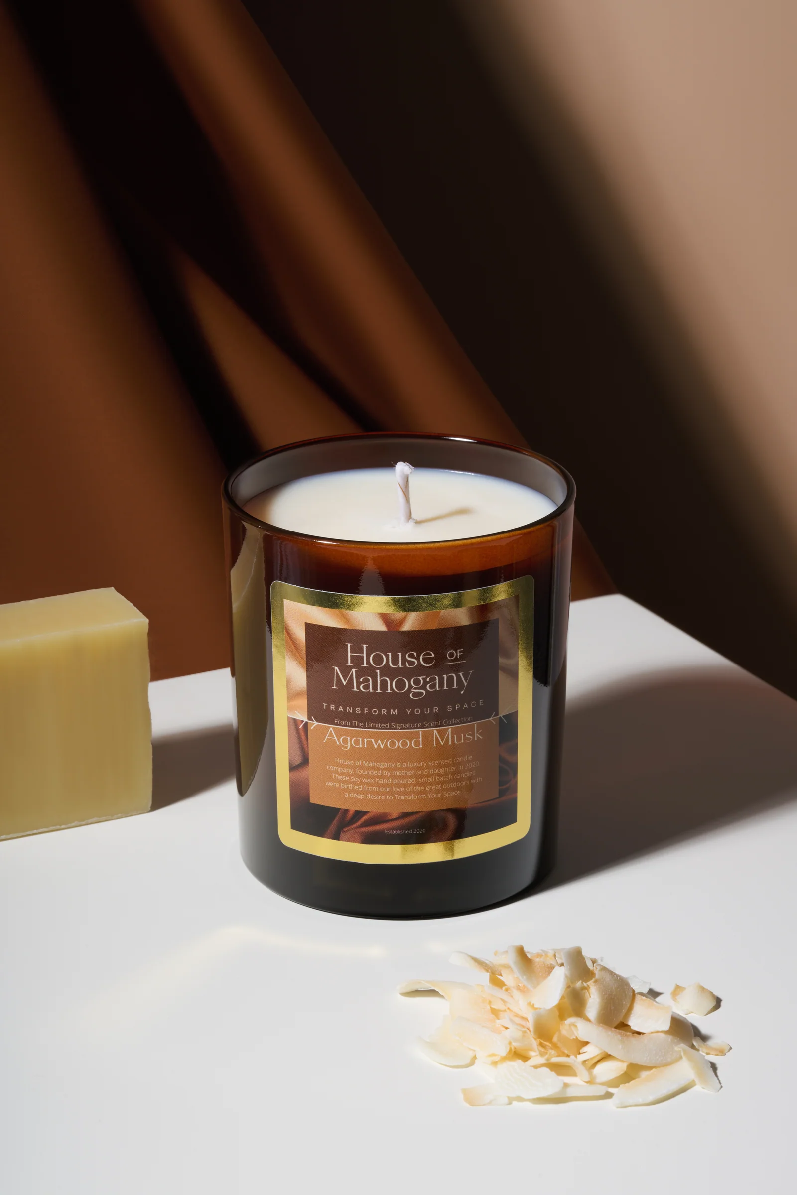 House of Mahogany candle in a dark glass vessel beside a yellow wax block and wax shavings against draped brown fabric.