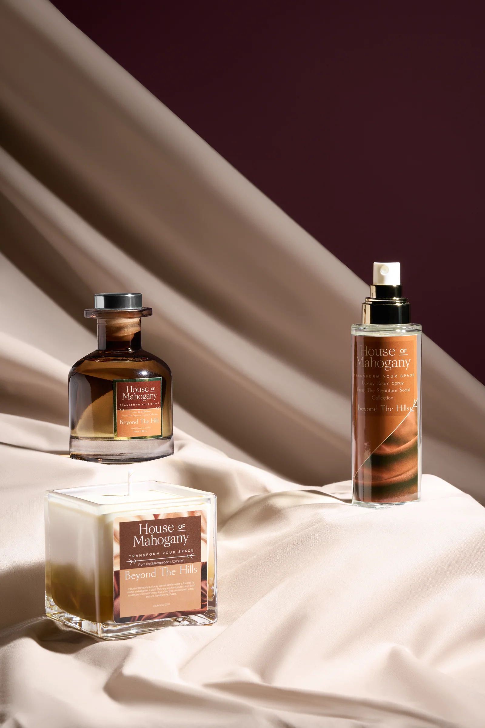 House of Mahogany fragrance bottle, room spray and candle arranged on cream fabric against burgundy.