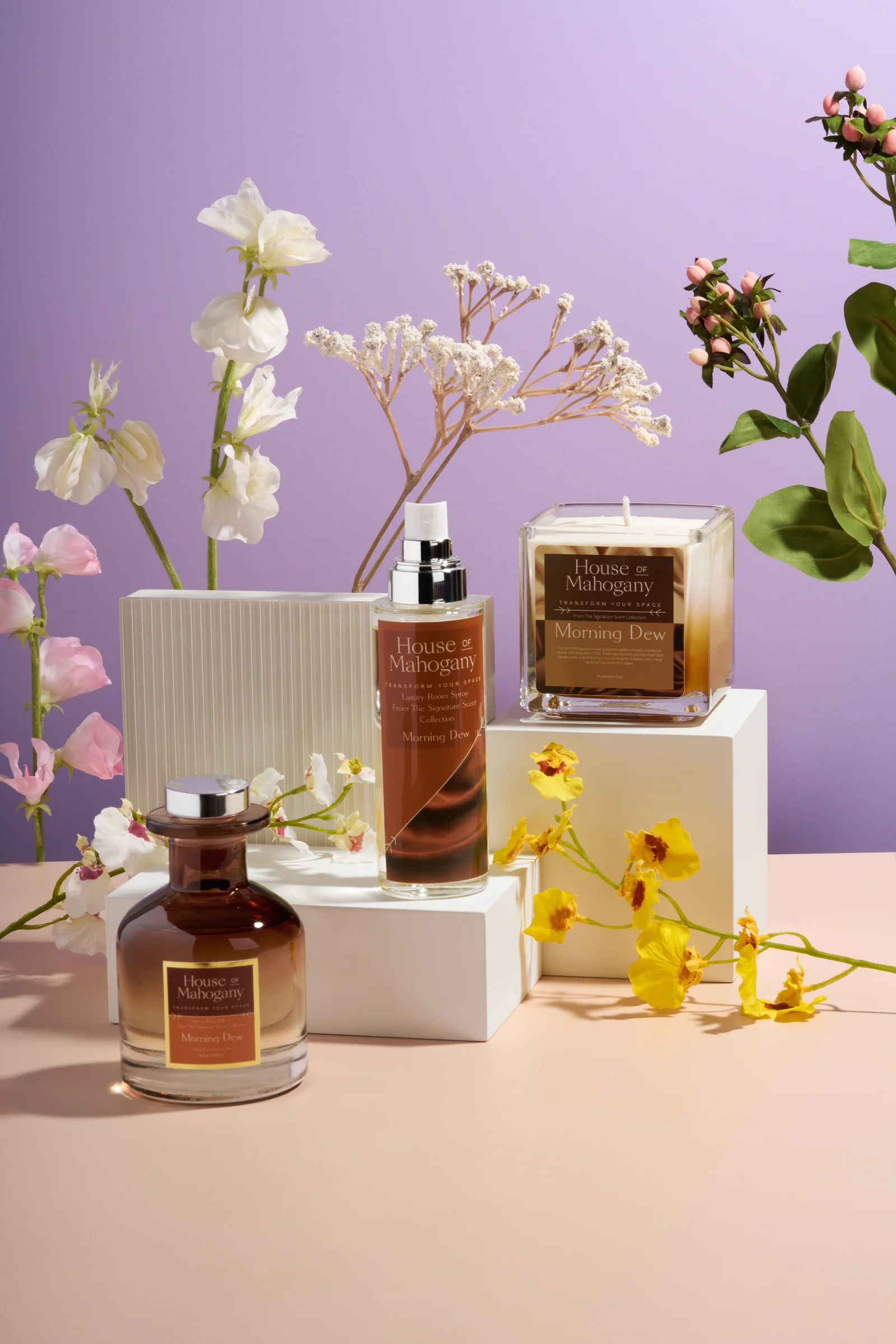House of Mahogany fragrance bottle, room spray and candle on white plinths with flowers against lilac.