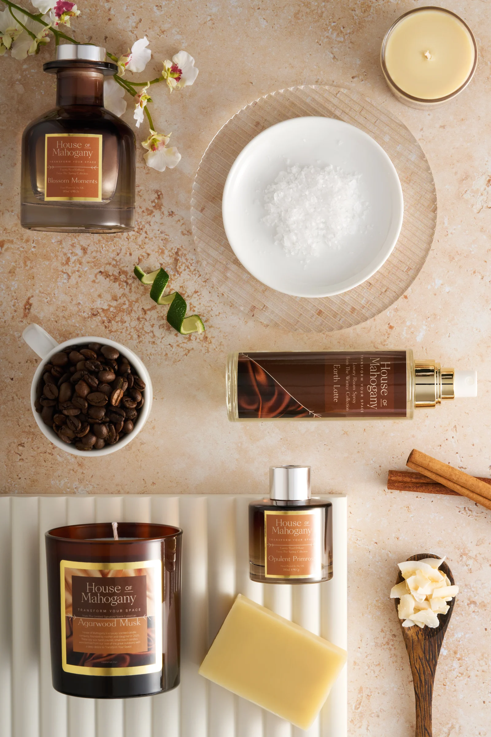 Overhead arrangement of House of Mahogany home fragrance products with flowers, coffee beans, cinnamon and wax on a beige surface.