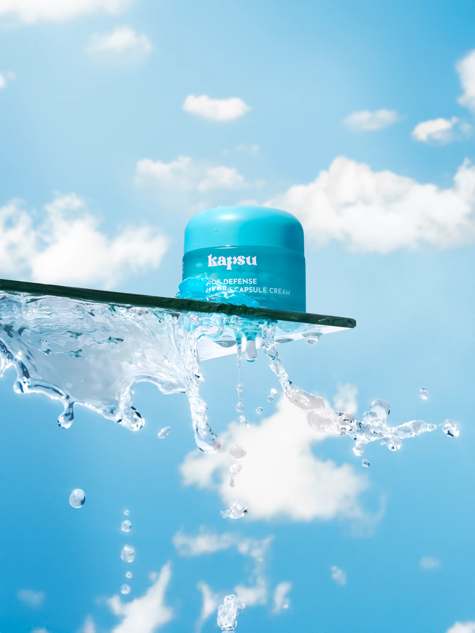 Turquoise Kapsu skincare jar on a glass shelf with splashing water against a blue sky.