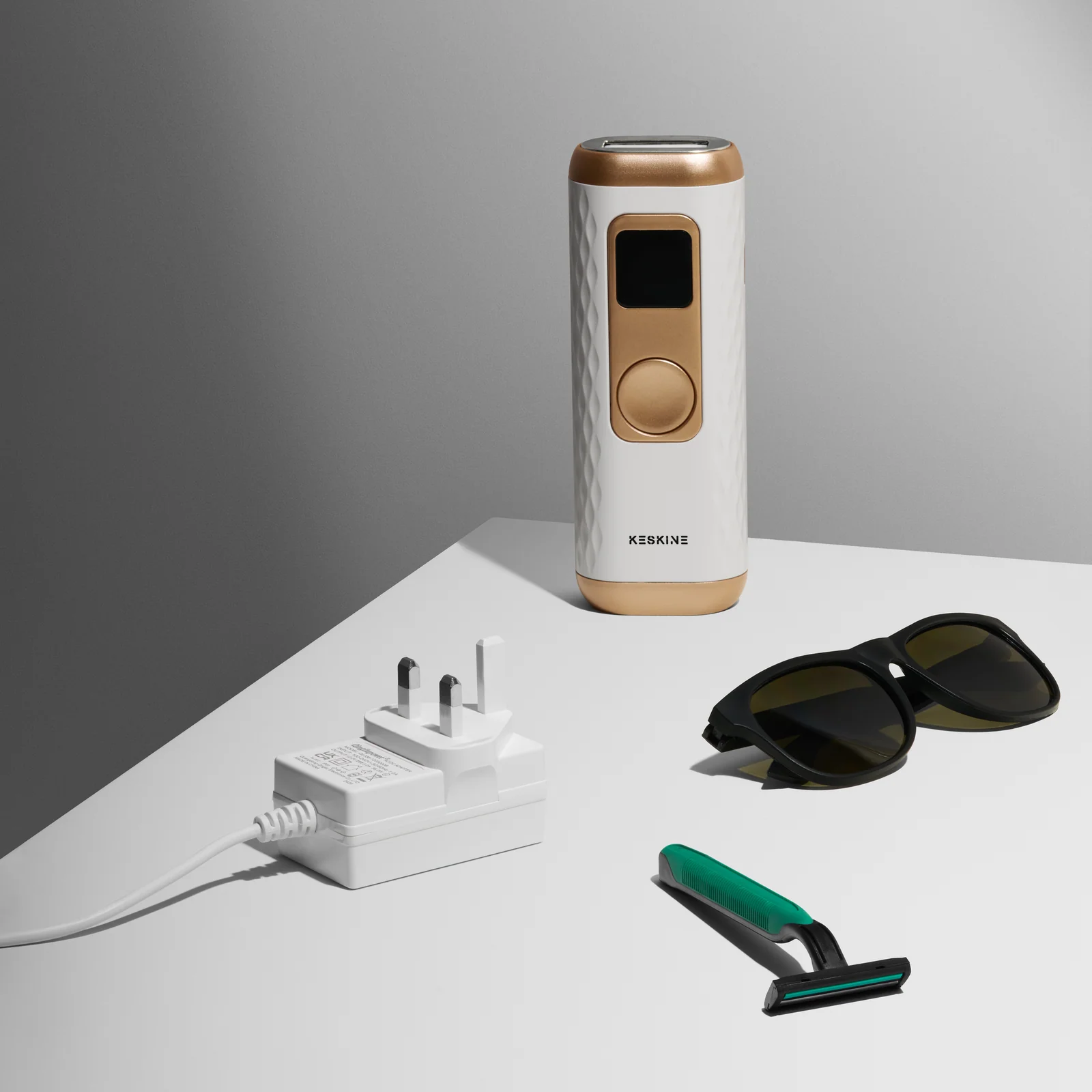 Keskine IPL device with a power adaptor, dark glasses and green razor on a grey platform.