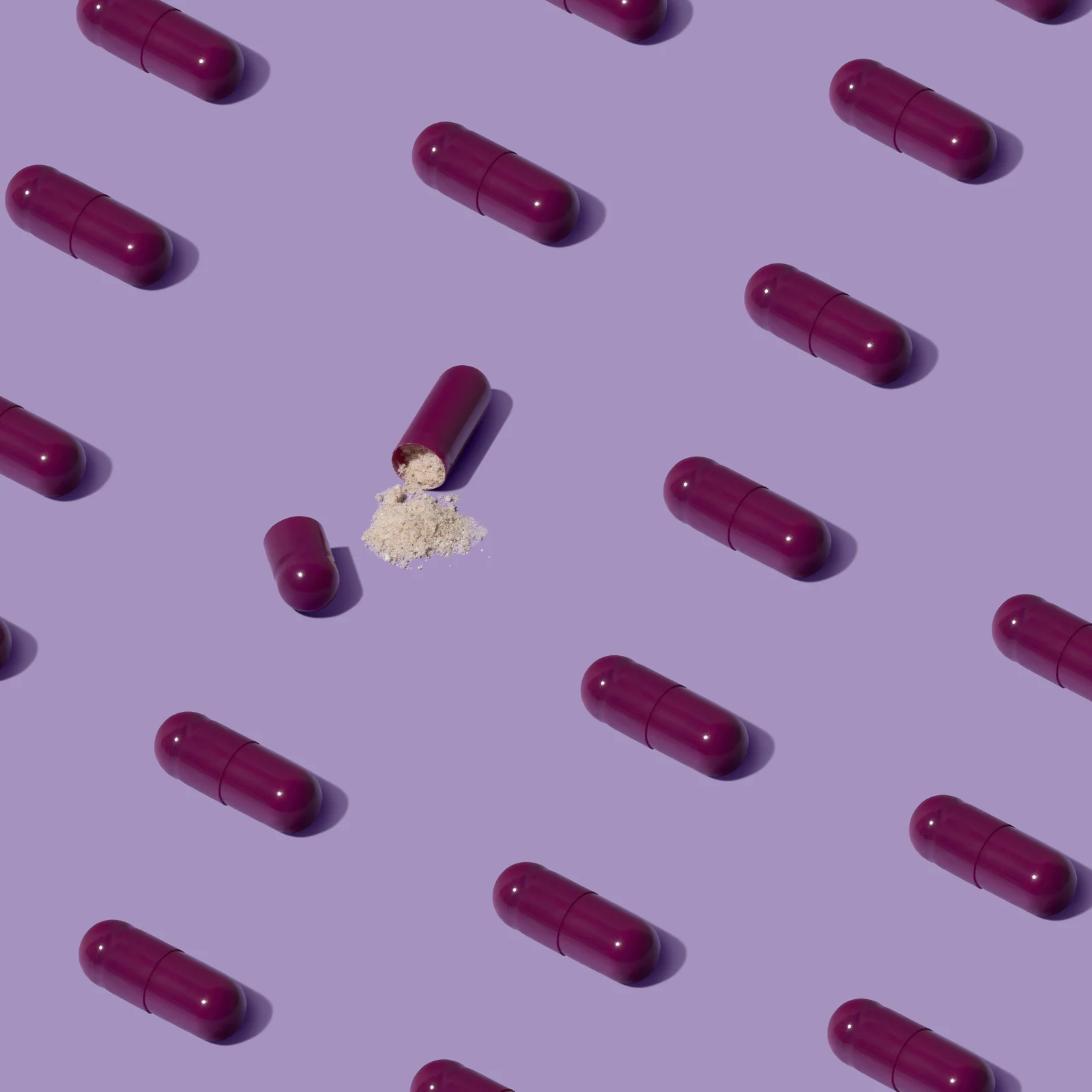 Purple supplement capsules arranged in a repeating pattern on lilac, with one open capsule spilling powder.