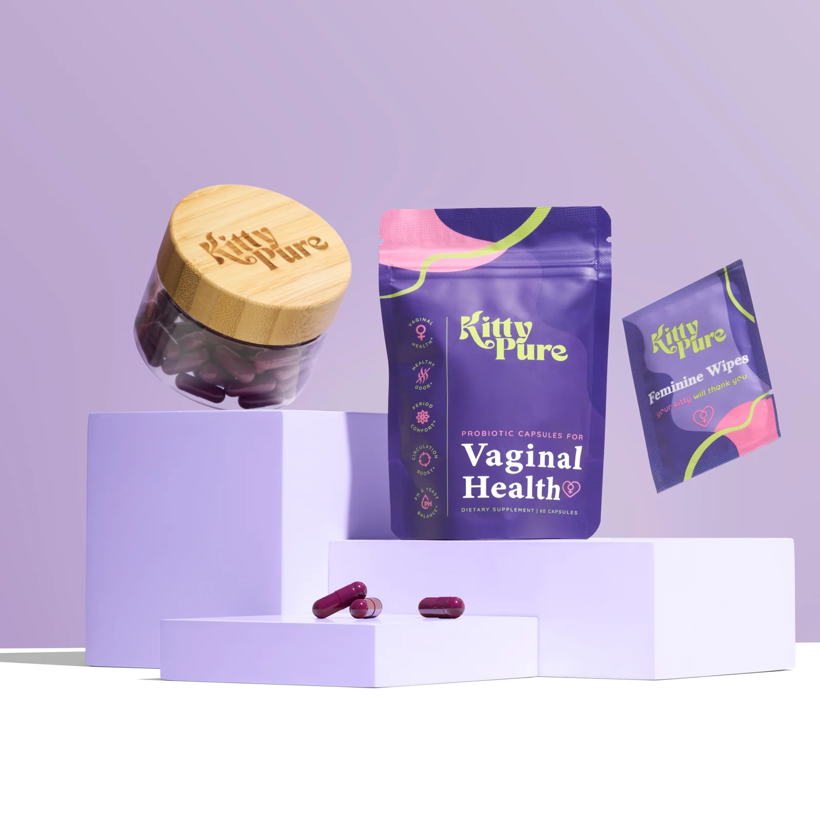 KittyPure supplement jar, Vaginal Health pouch and wipes sachet on lilac plinths with loose capsules.