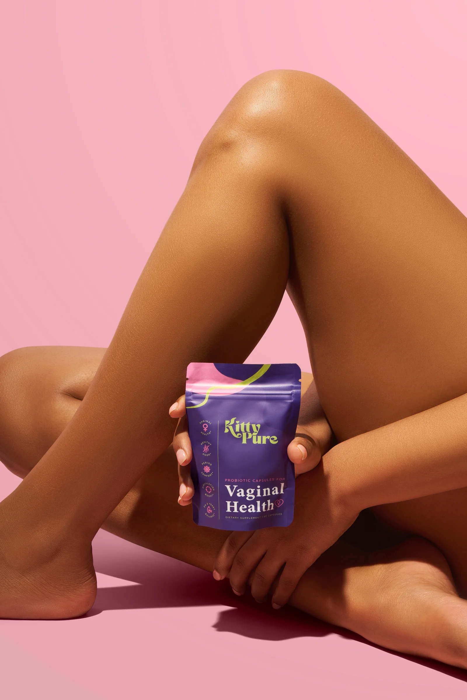 Seated person holding a KittyPure Vaginal Health supplement pouch beside a bent leg on a pink set.