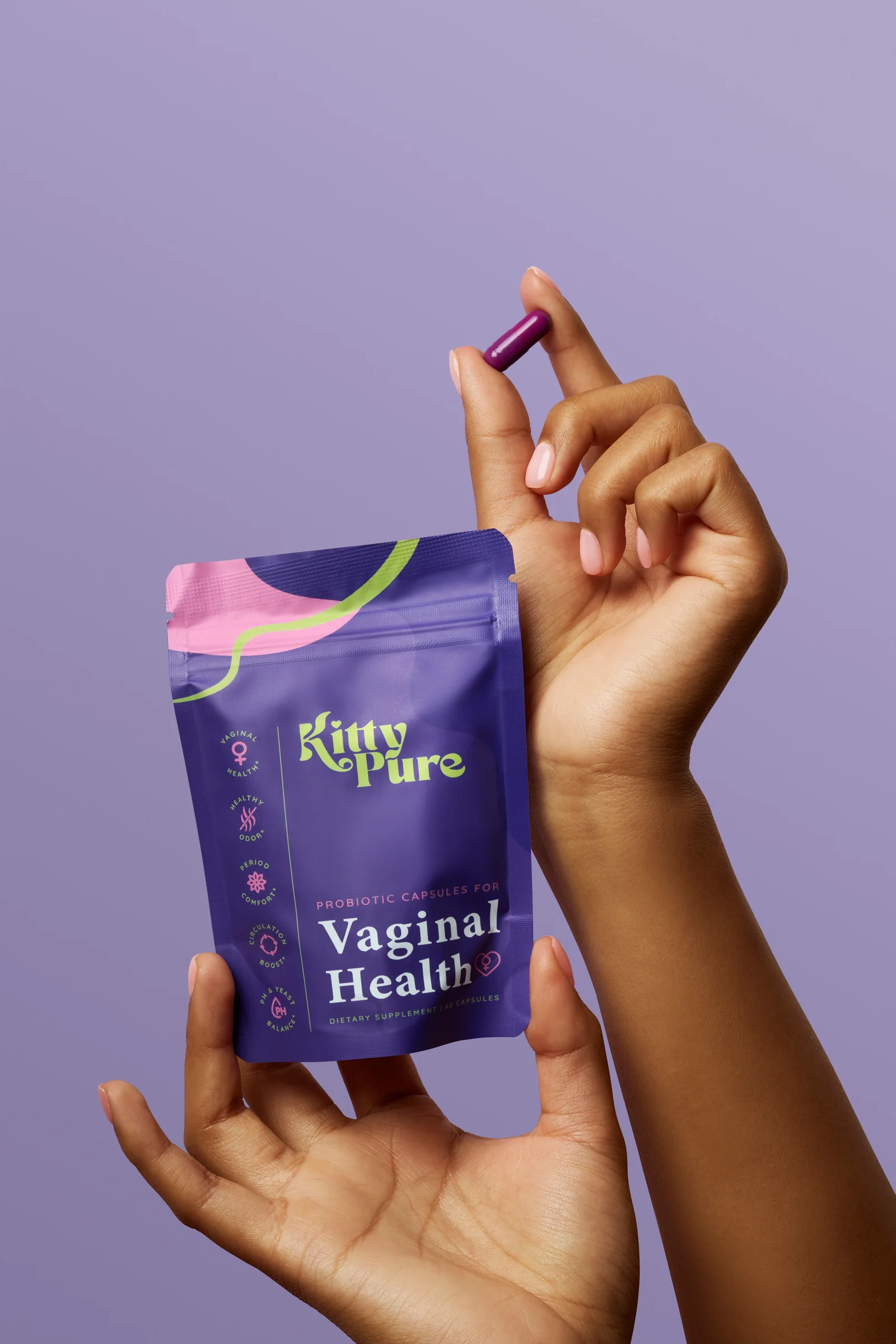 Hands displaying a KittyPure Vaginal Health supplement pouch and a single purple capsule against lilac.