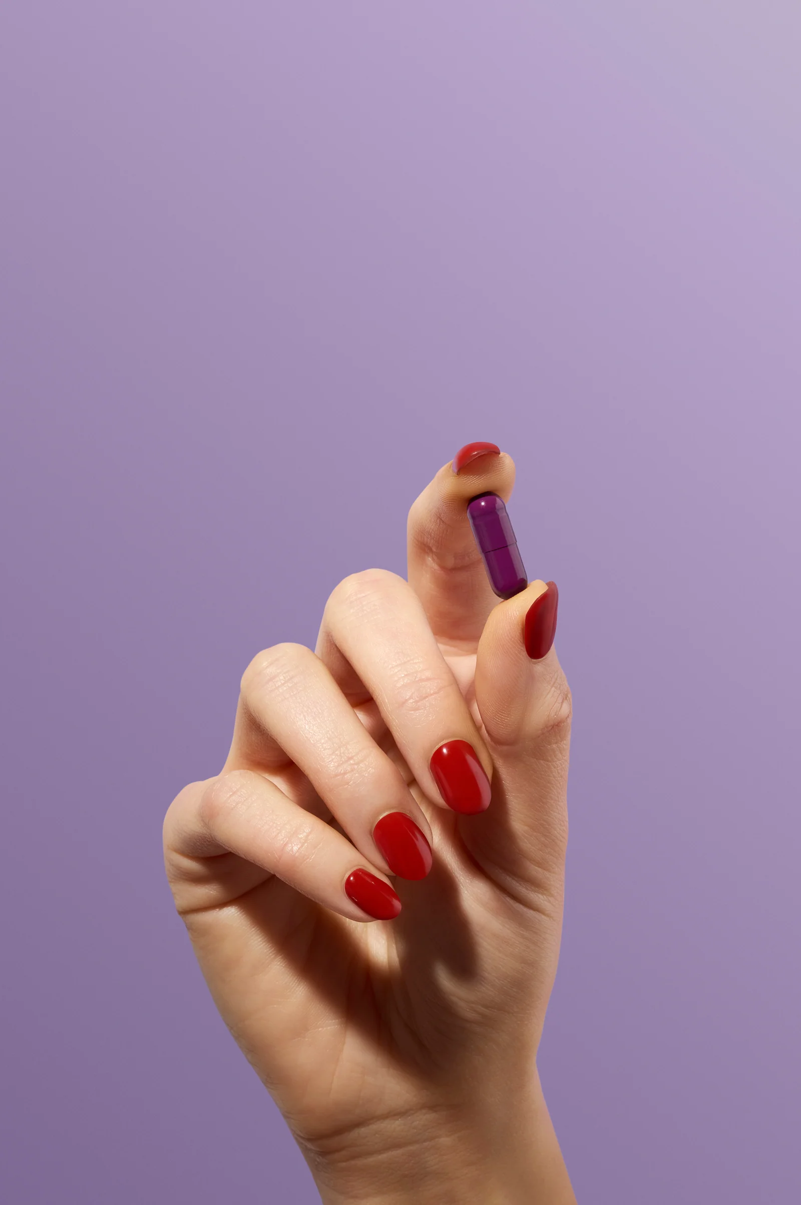 Hand with red-painted nails holding a purple supplement capsule between finger and thumb against lilac.