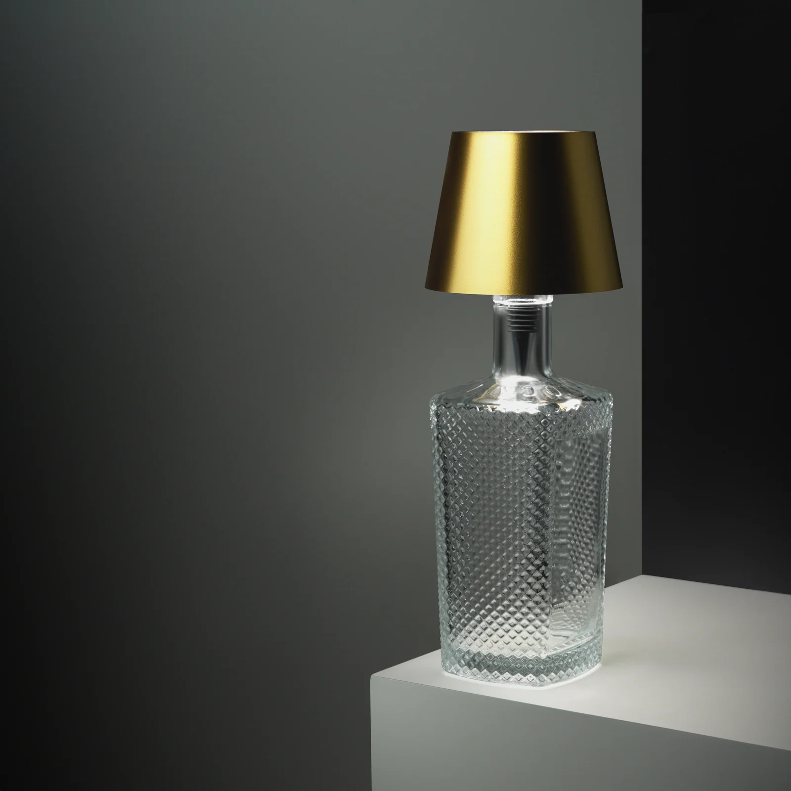 Gold Liquor Lit lamp top fitted to a clear textured bottle on a grey plinth.