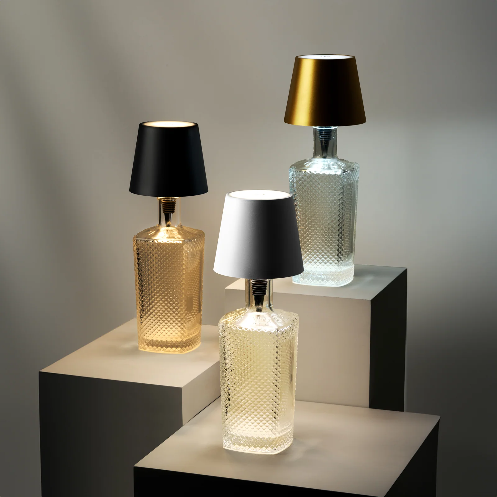 Three textured bottle lamps with black, silver and gold Liquor Lit tops arranged on stepped neutral plinths.