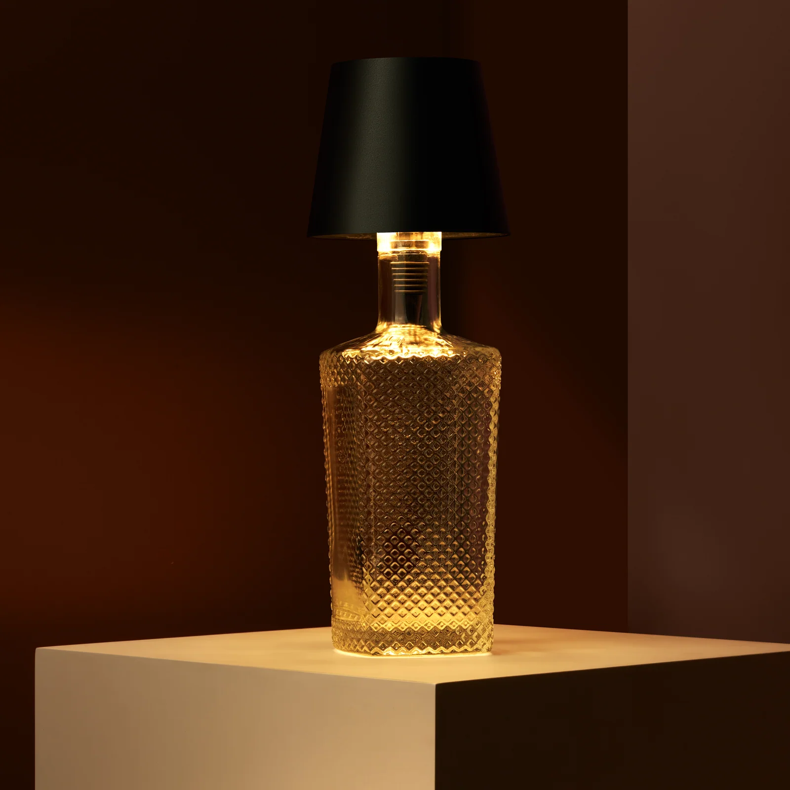 Glowing bottle lamp with a black Liquor Lit top on a cream plinth against dark brown.