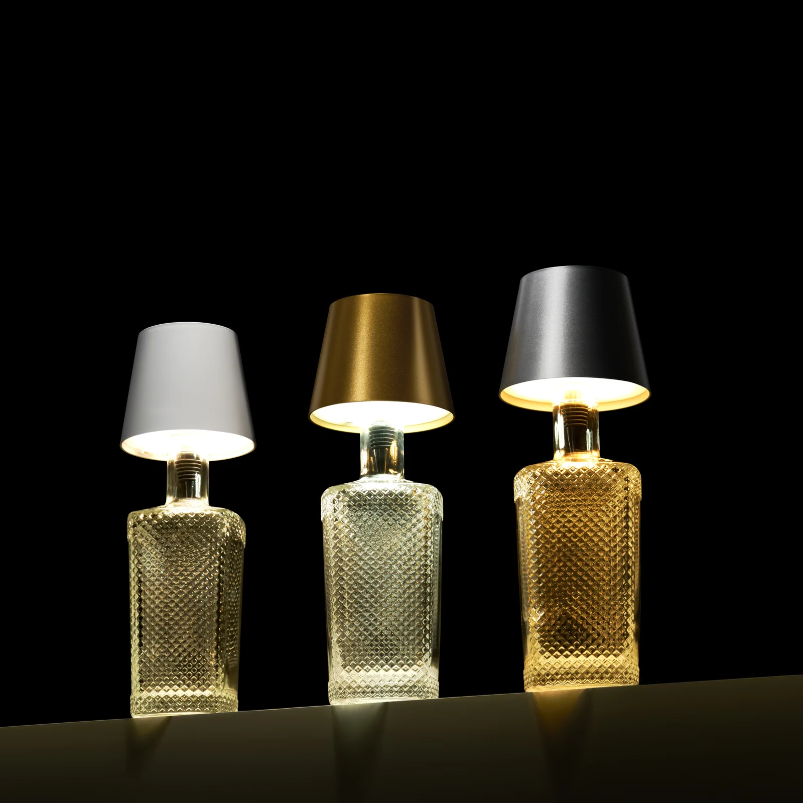 Three illuminated bottle lamps with white, gold and silver Liquor Lit tops against black.