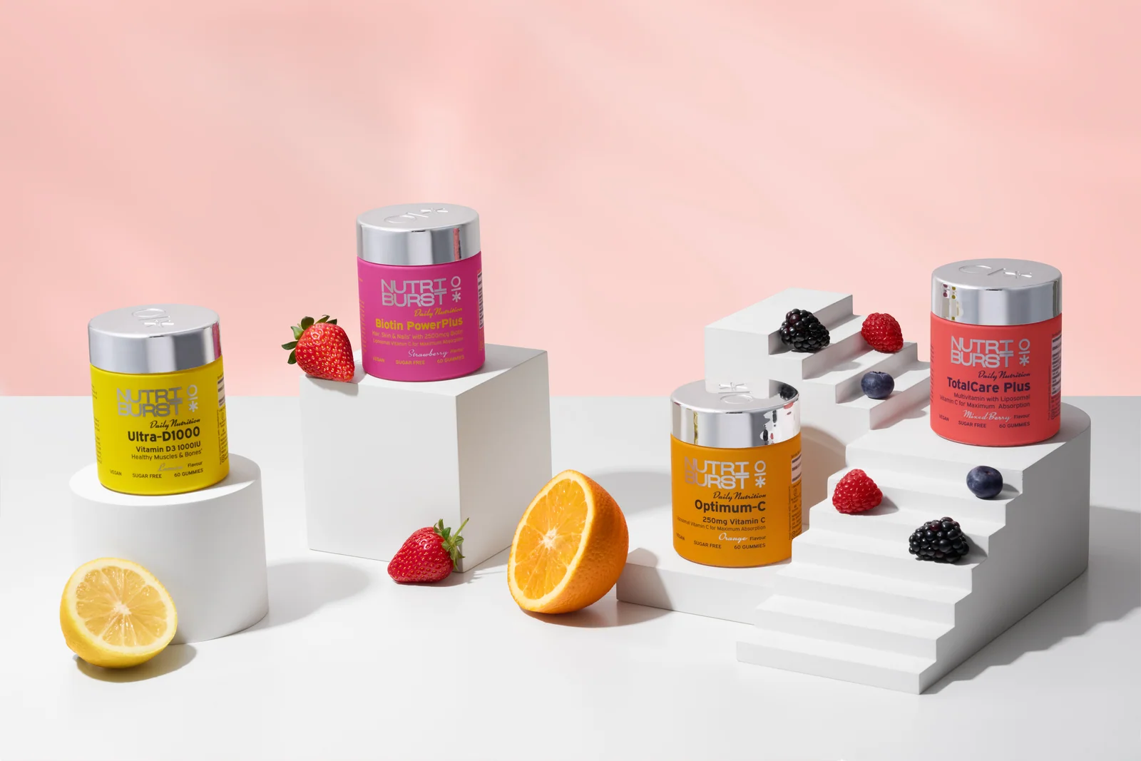 Four colourful Nutriburst jars with citrus fruit and berries on white geometric platforms against pale pink.