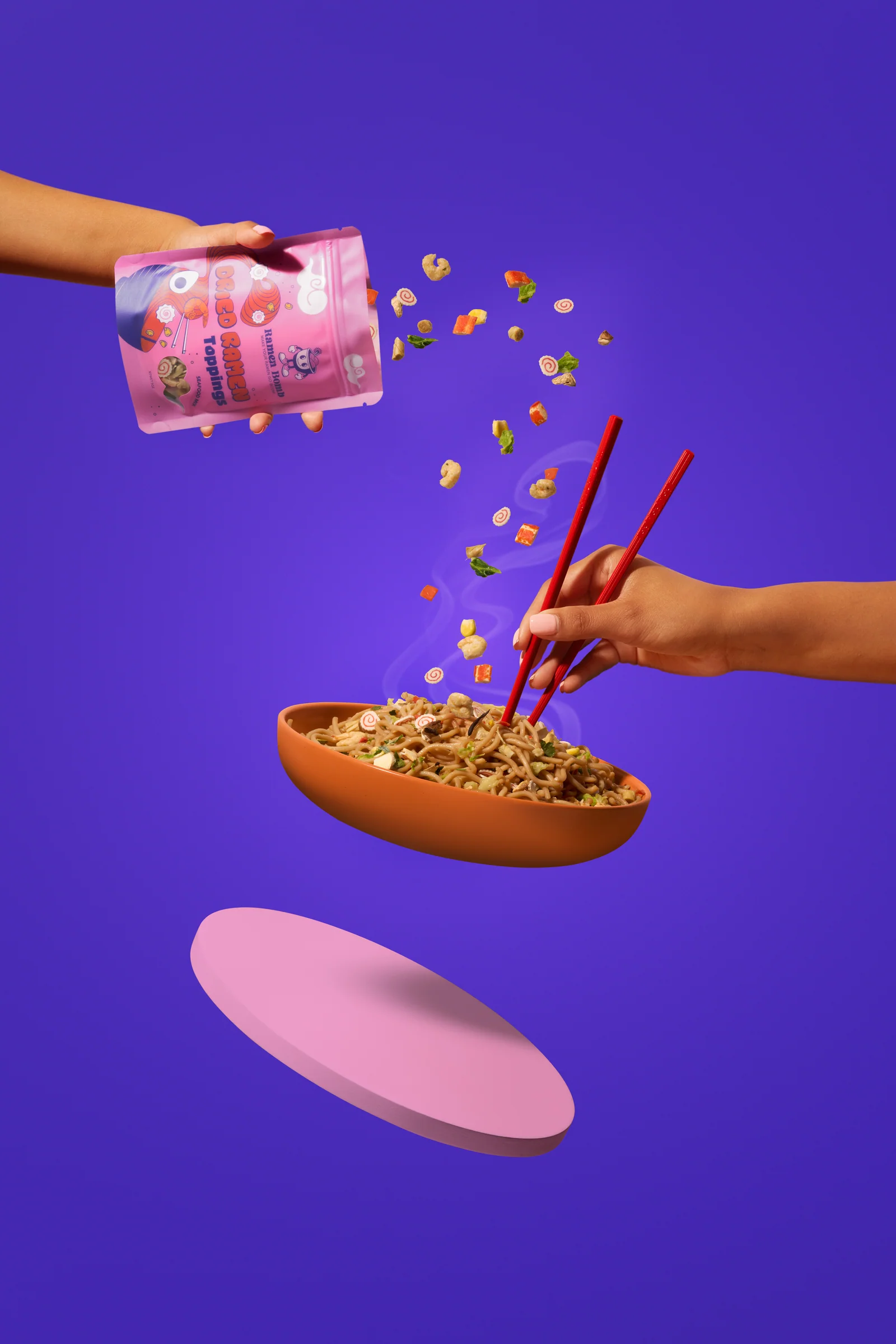 Hands pouring Ramen Bomb toppings towards a bowl of noodles against purple.