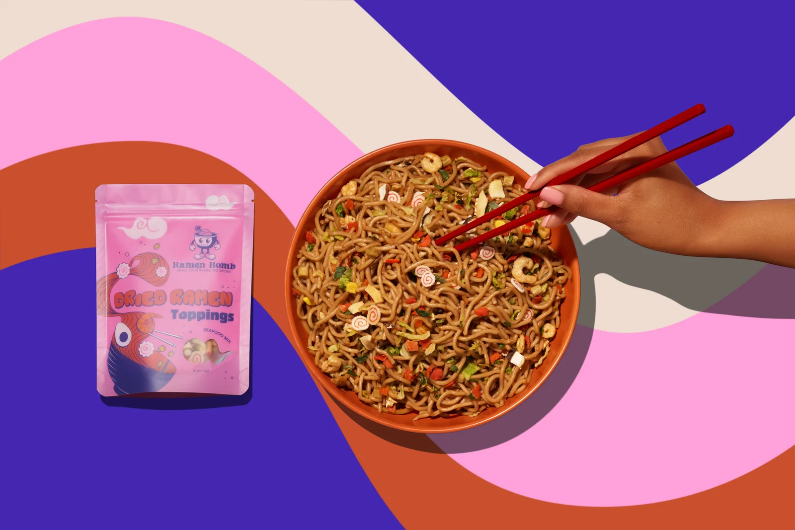 Overhead view of a Ramen Bomb packet beside a bowl of noodles and a hand holding chopsticks on a colourful patterned surface.