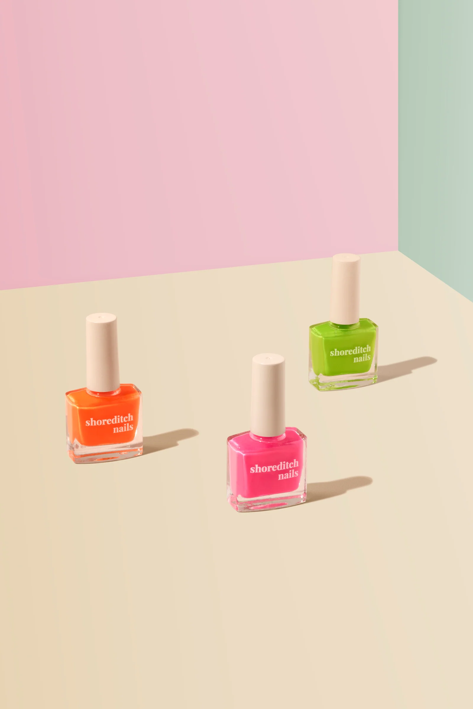 Orange, pink and green Shoreditch Nails polish bottles casting shadows on a cream surface against pastel pink and green.