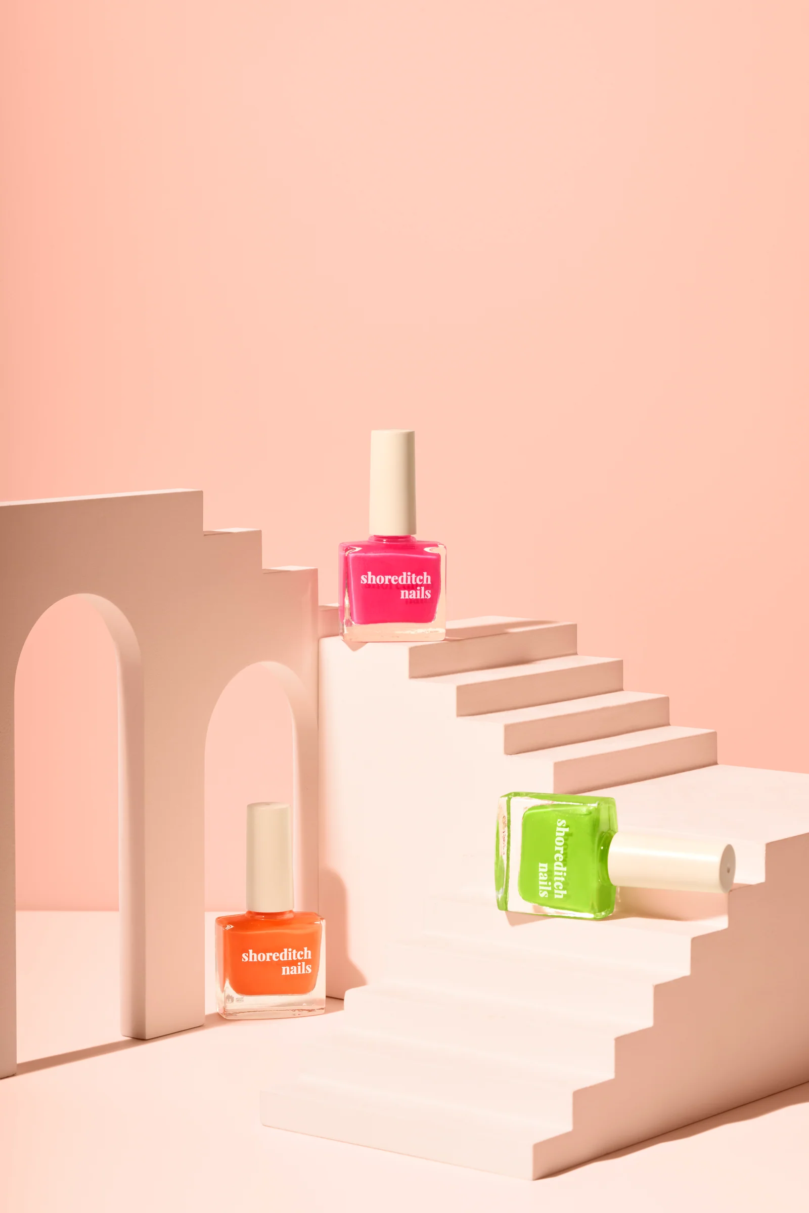 Pink, green and orange Shoreditch Nails polish bottles on a pale pink staircase beside an arch against peach.