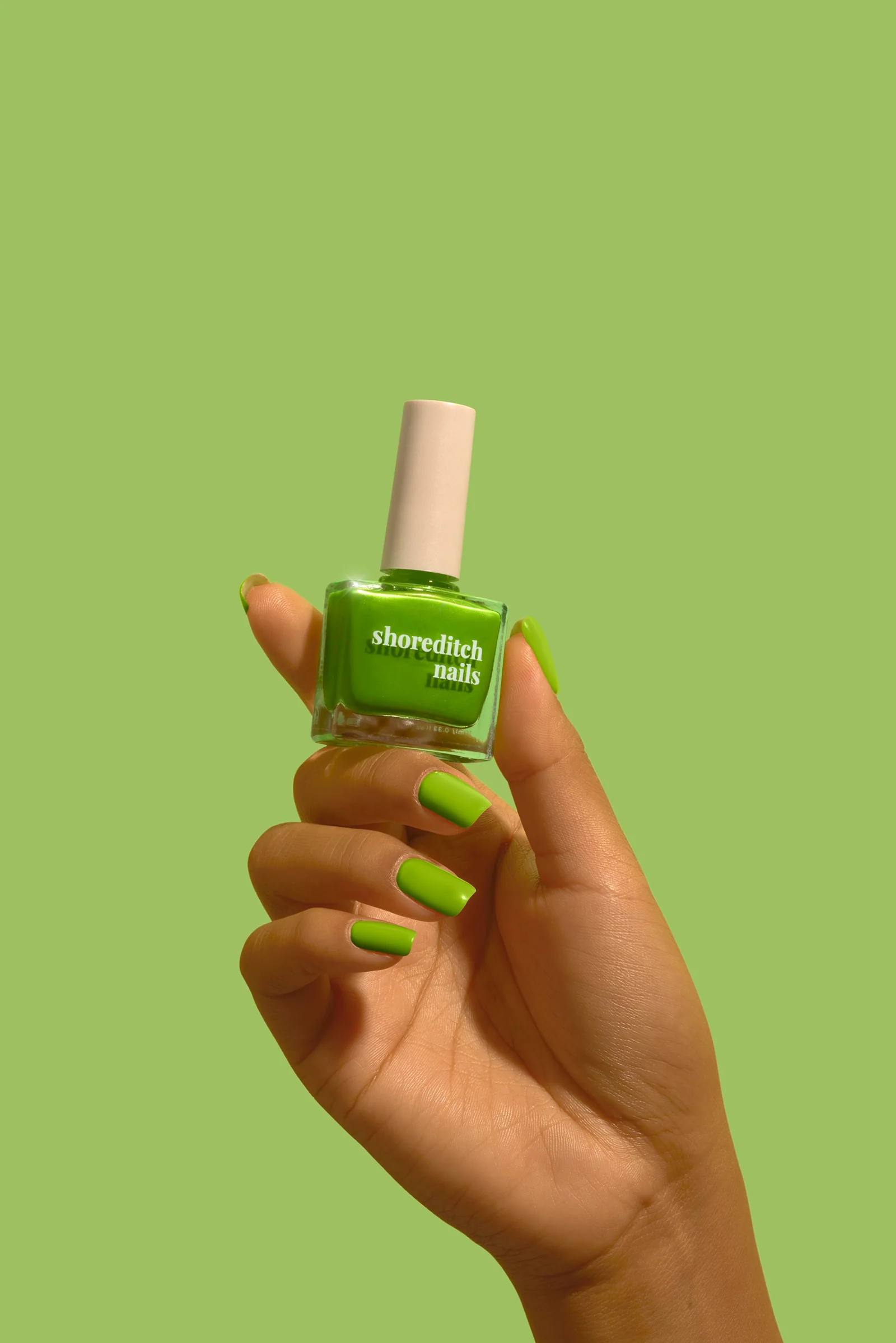 Hand with green-painted nails holding a green Shoreditch Nails polish bottle against a matching green background.